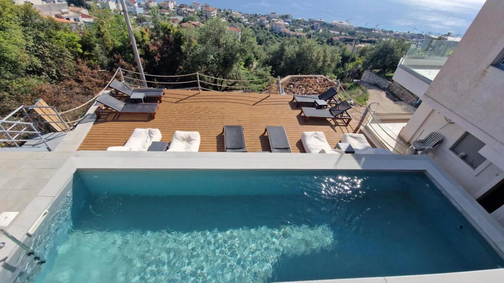 Looking down over the pool at Ionian Pearl to the timber deck with loungers and bean bags, and beyond it the olive slope, the village roofs and the sea