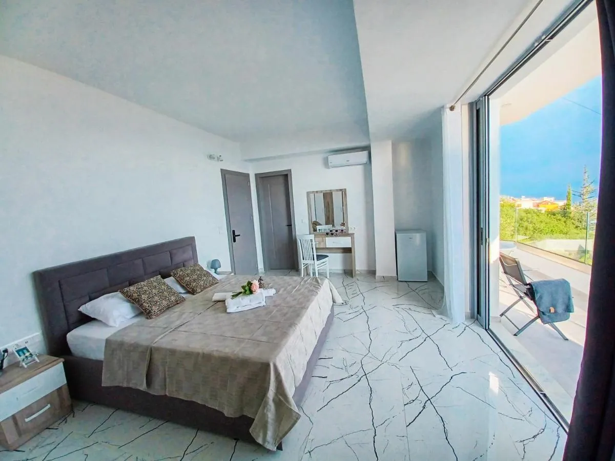 A double bed with a grey headboard in a white room at Ionian Pearl, a dressing table and a full-height glass door to a balcony with a glimpse of the sea