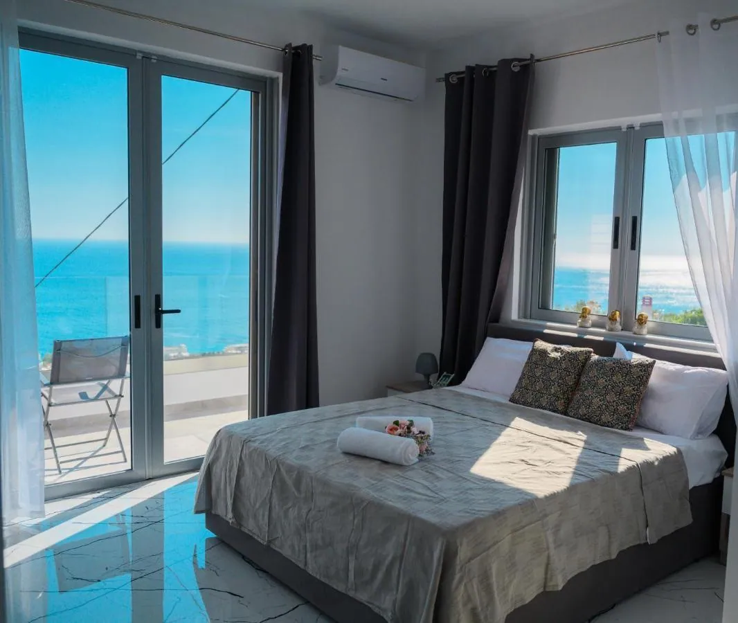 A double bed made up with towels in a bright room at Ionian Pearl, with sheer curtains, a glass door to the balcony and the sea beyond