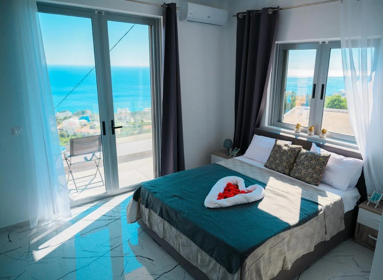 A double bed with a teal throw in a white room at Ionian Pearl, its glass doors and window open onto a balcony and the sea