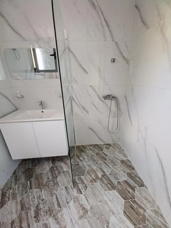 A shower room at Ionian Pearl tiled in white marble-effect porcelain with a glass screen, a basin unit and a wood-effect floor