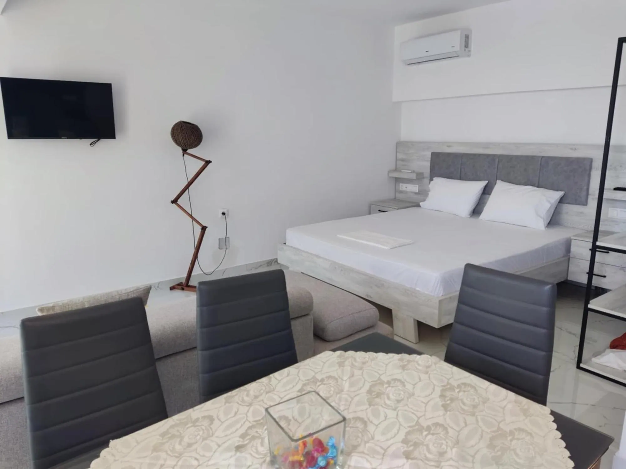 A white room at Ionian Pearl with a double bed, a dining table with four grey chairs, a wall-mounted TV and an open clothes rail