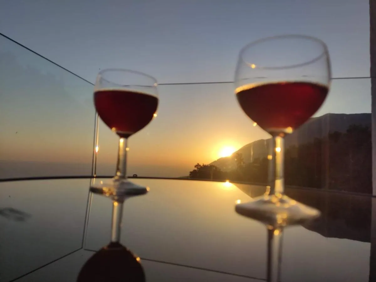 Two glasses of red wine on a glass table at Ionian Pearl, with the sun setting over the sea behind a headland
