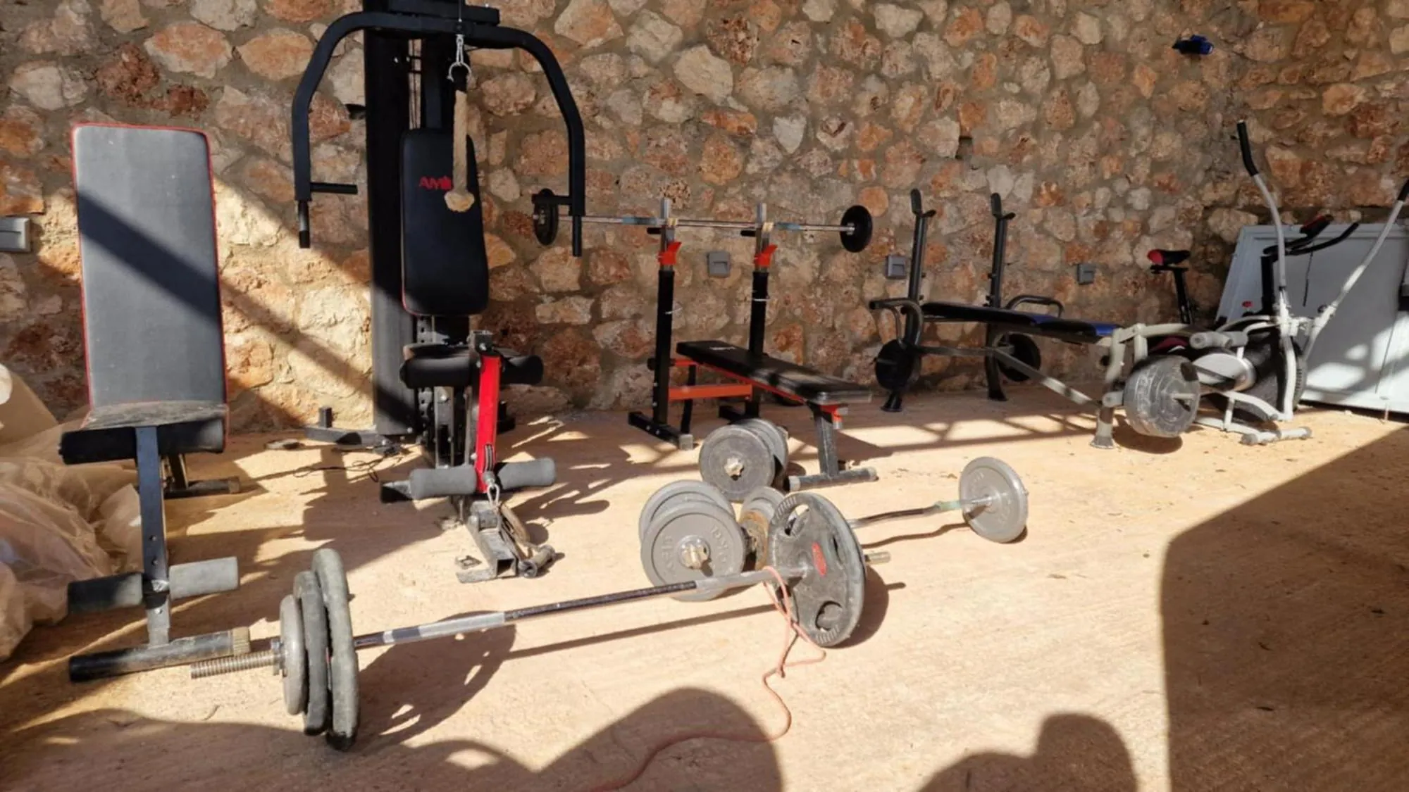 A multi-gym, a bench and a rack of free weights standing on a stone floor against a drystone wall at Ionian Pearl