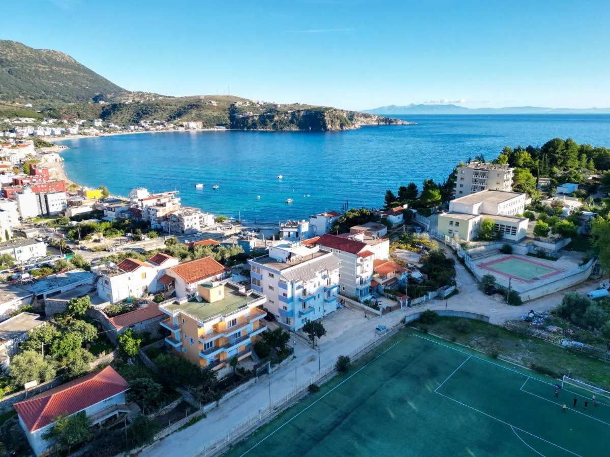 Aerial photograph looking south over the bay at Himarë, with the white and blue apartment building beside an orange one, the corner of the football pitch below them, and the headland and open sea beyond