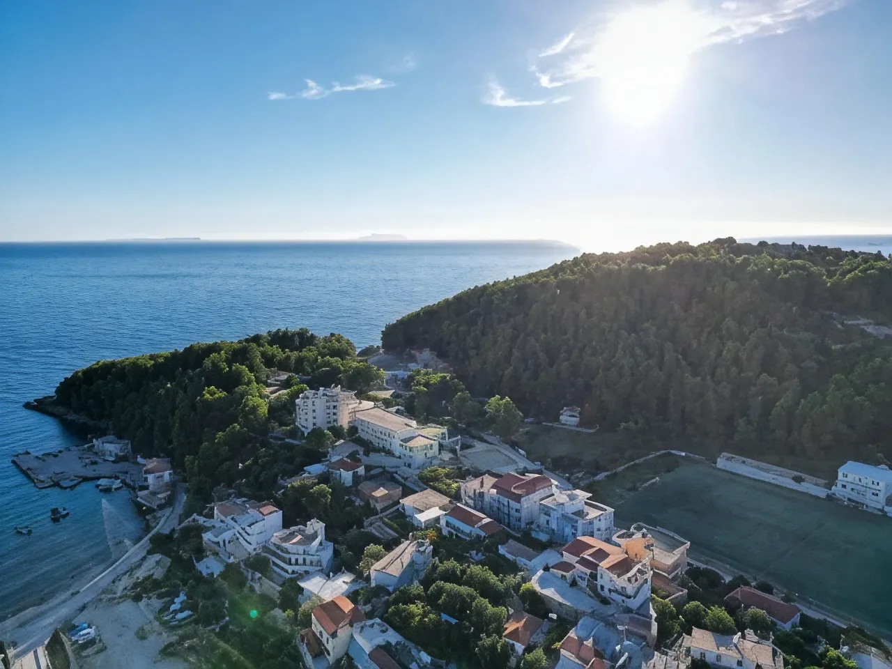 Aerial photograph in low evening sun over the wooded headland at Himarë, the small harbour on its left, white houses below and the pitch at the right edge