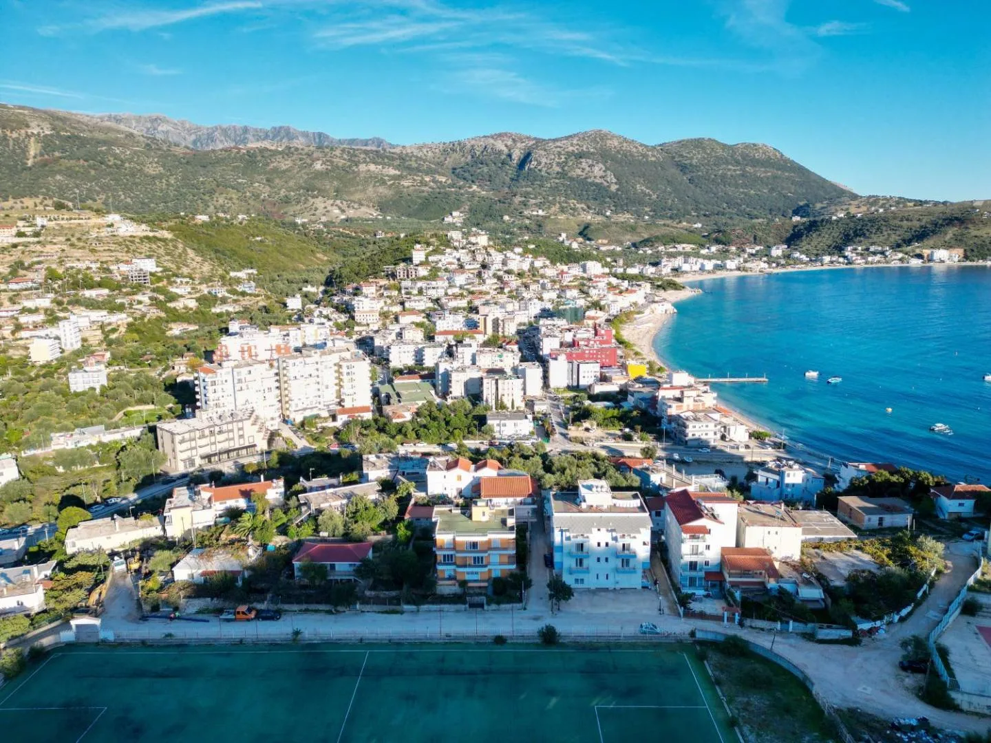 Aerial photograph of Himarë: the green artificial-turf pitch of the Petro Ruci Stadium in the foreground, the white apartment building with blue balconies across the road from it, and the town and the curve of Spile Beach running down to the bay beyond