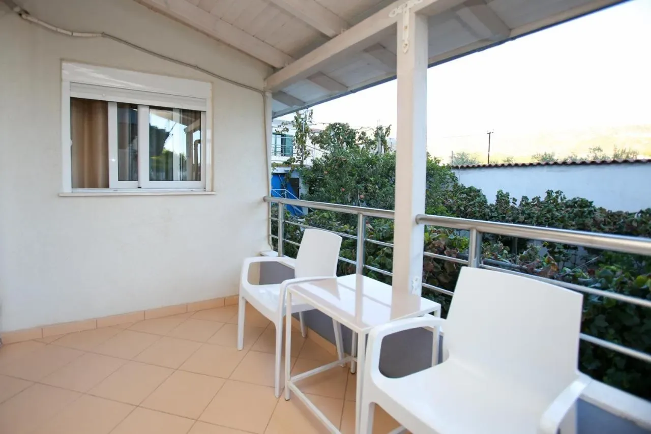 A covered balcony with two white chairs and a small table, tiled floor, the steel rail opening onto a garden of vines and fruit trees