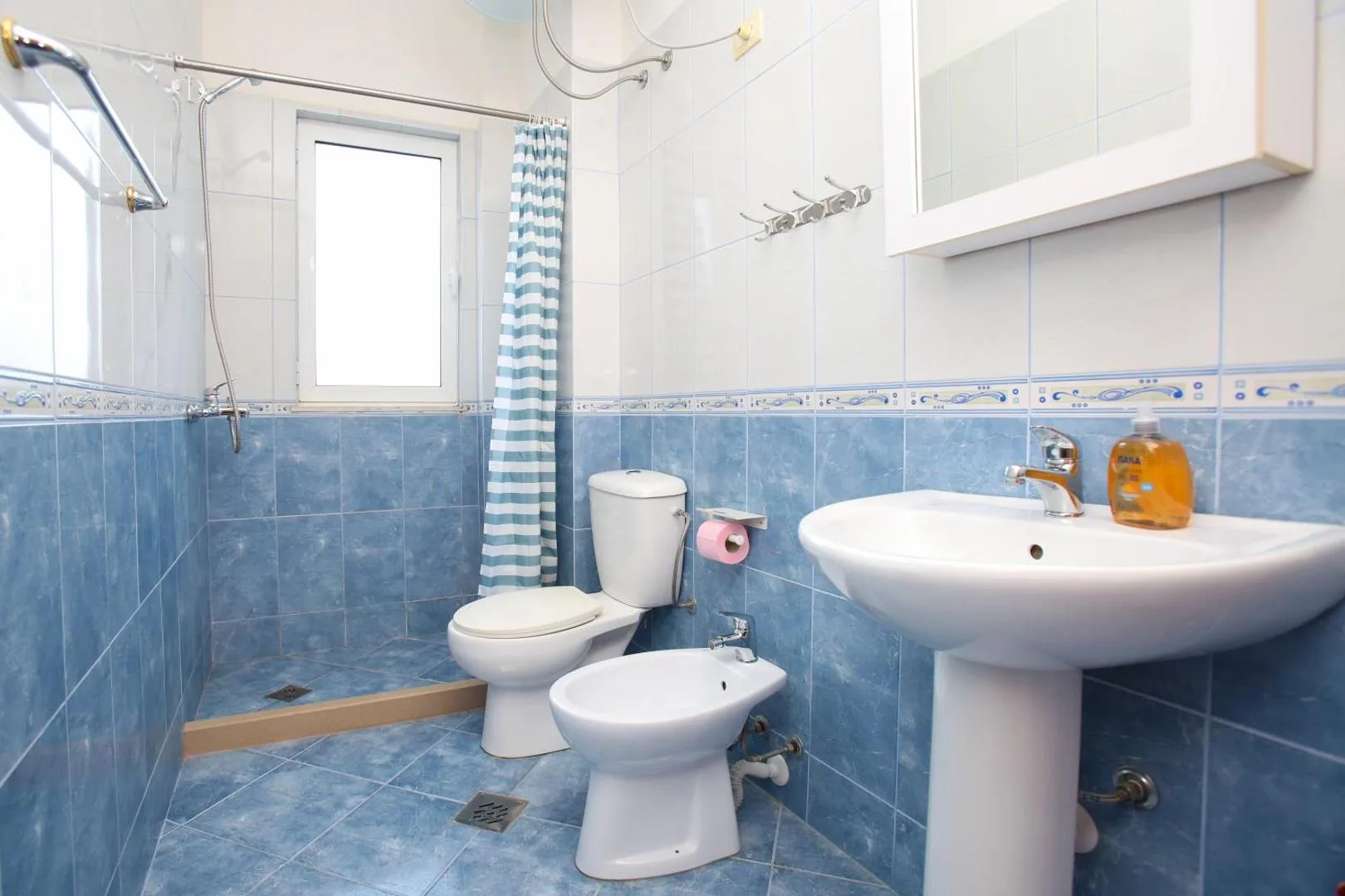 A bathroom tiled in blue to dado height and white above, with a walk-in shower behind a striped curtain, a basin, a toilet and a bidet