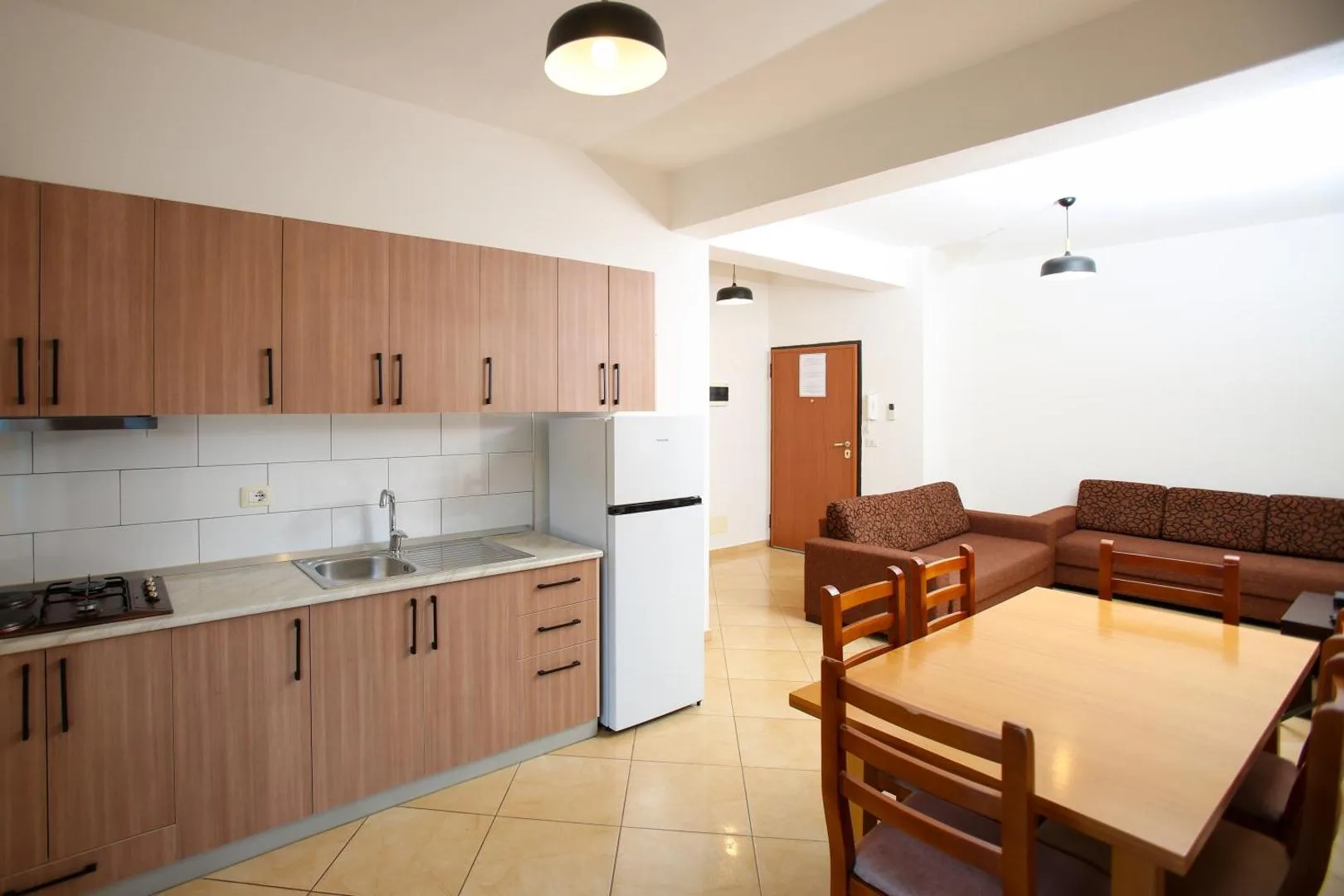 An open-plan apartment: a wood-fronted kitchen with a gas hob, sink and tall fridge, a dining table with four wooden chairs and two brown sofas by the front door