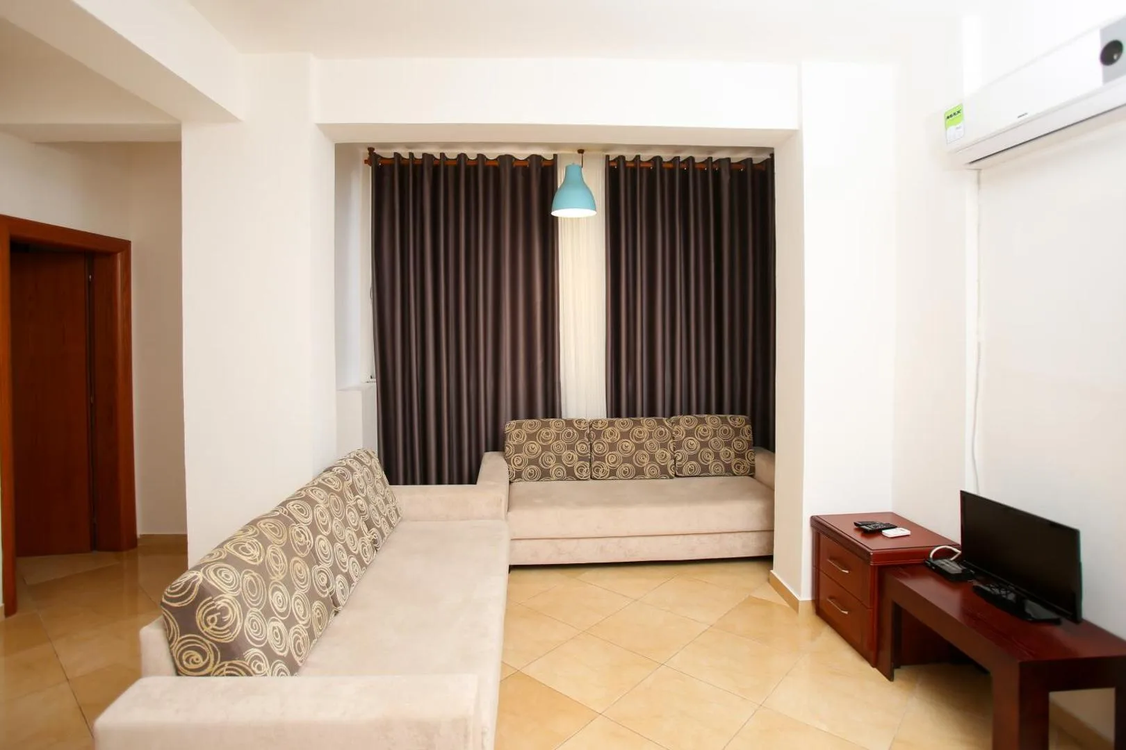 A living room with a cream corner sofa in an alcove under dark curtains, a television on a cherry-wood stand and an air-conditioning unit high on the wall