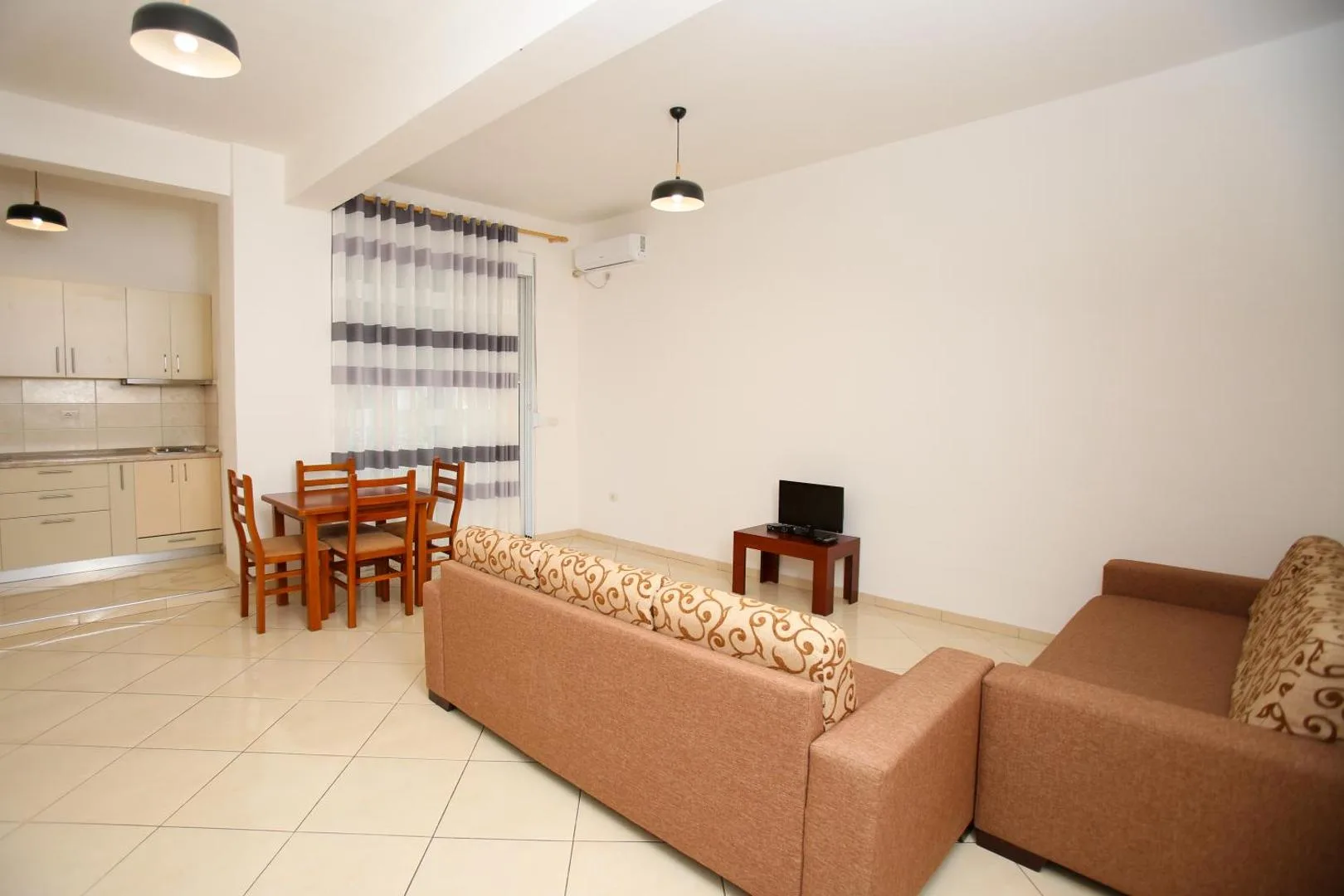 A bright living room with two sofas, a small television and a dining table for four by the kitchen, a striped curtain over the balcony door