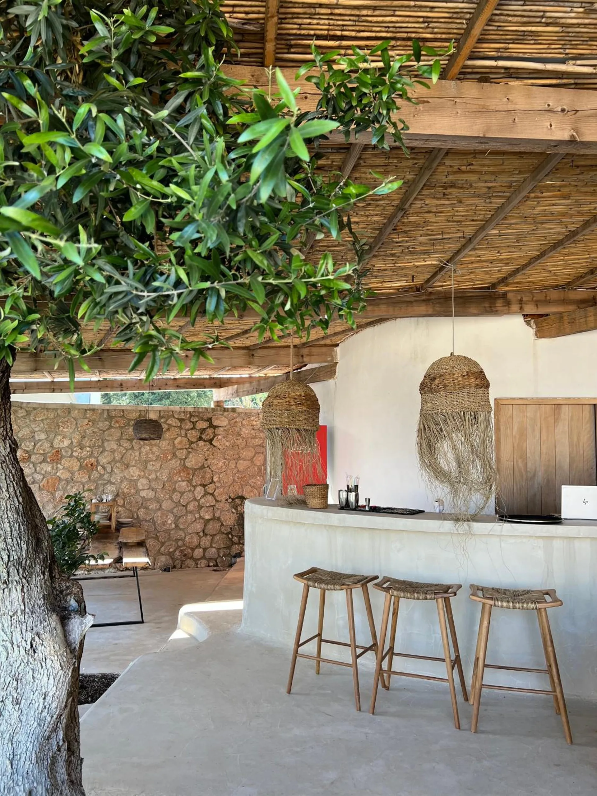 A white microcement bar with three wooden stools under the reed roof, an olive branch hanging into the frame and a jute pendant over the counter