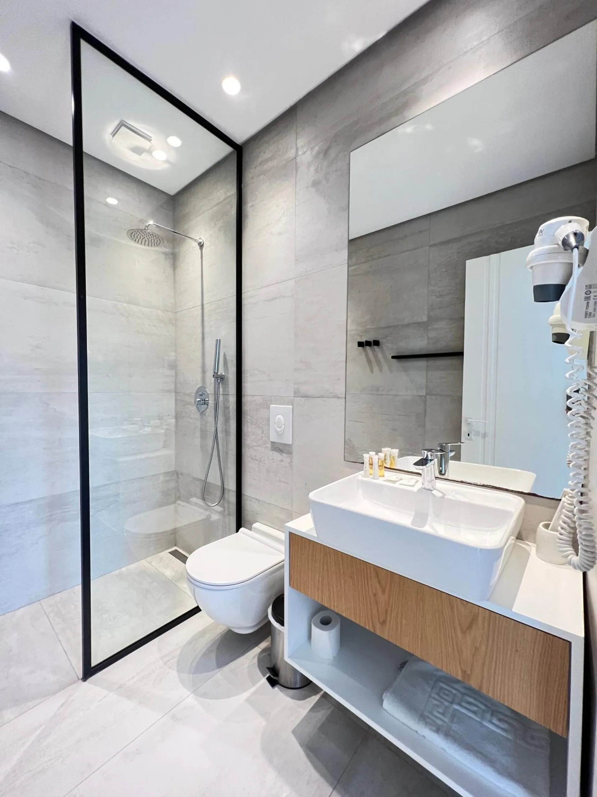 A grey-tiled bathroom with a glass walk-in shower, a white basin on a walnut vanity and a wall-mounted hairdryer