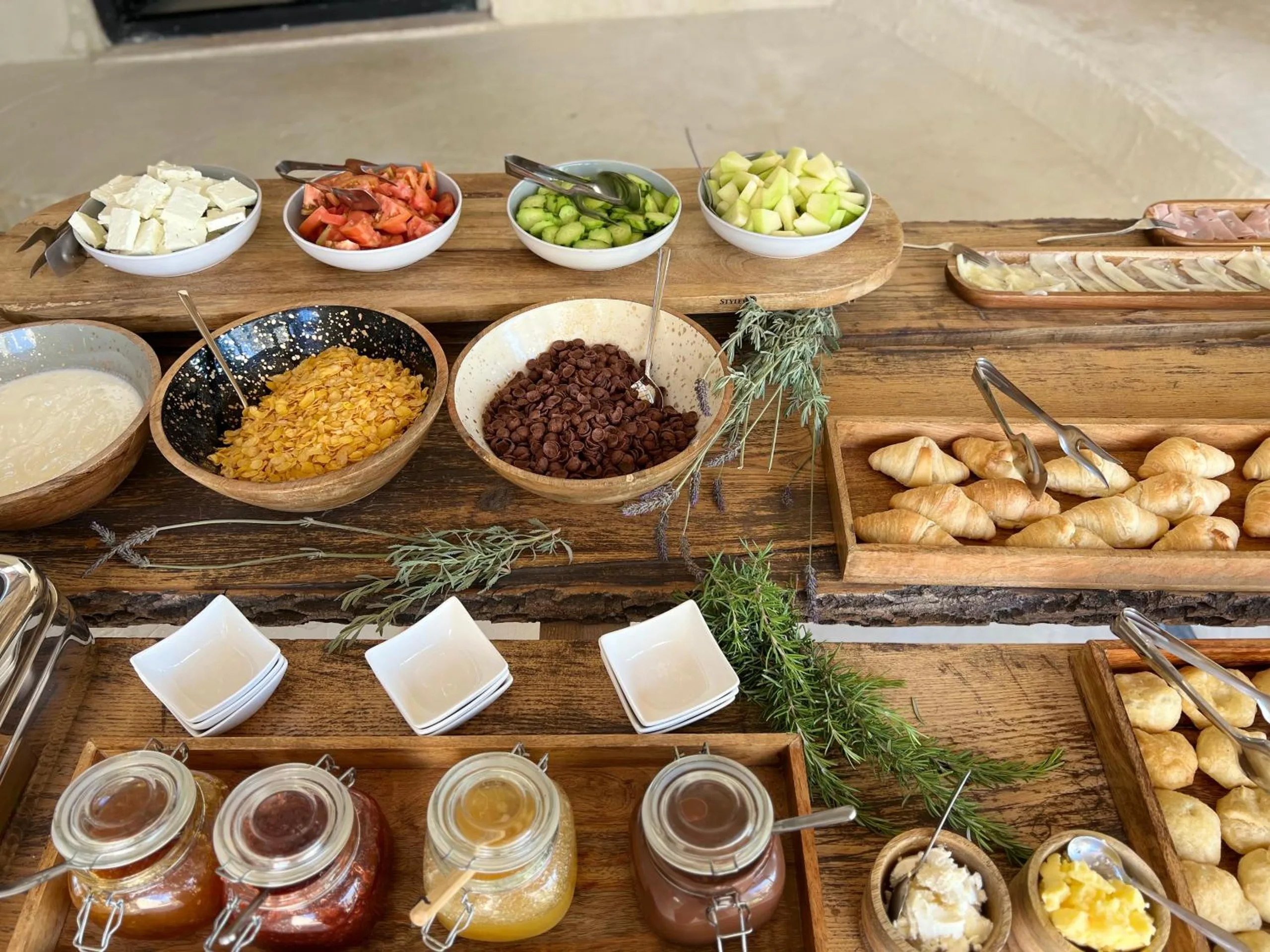 Close view of the breakfast boards: white cheese, sliced tomato, cucumber, olives, cereals, croissants and jars of honey and jam with sprigs of rosemary