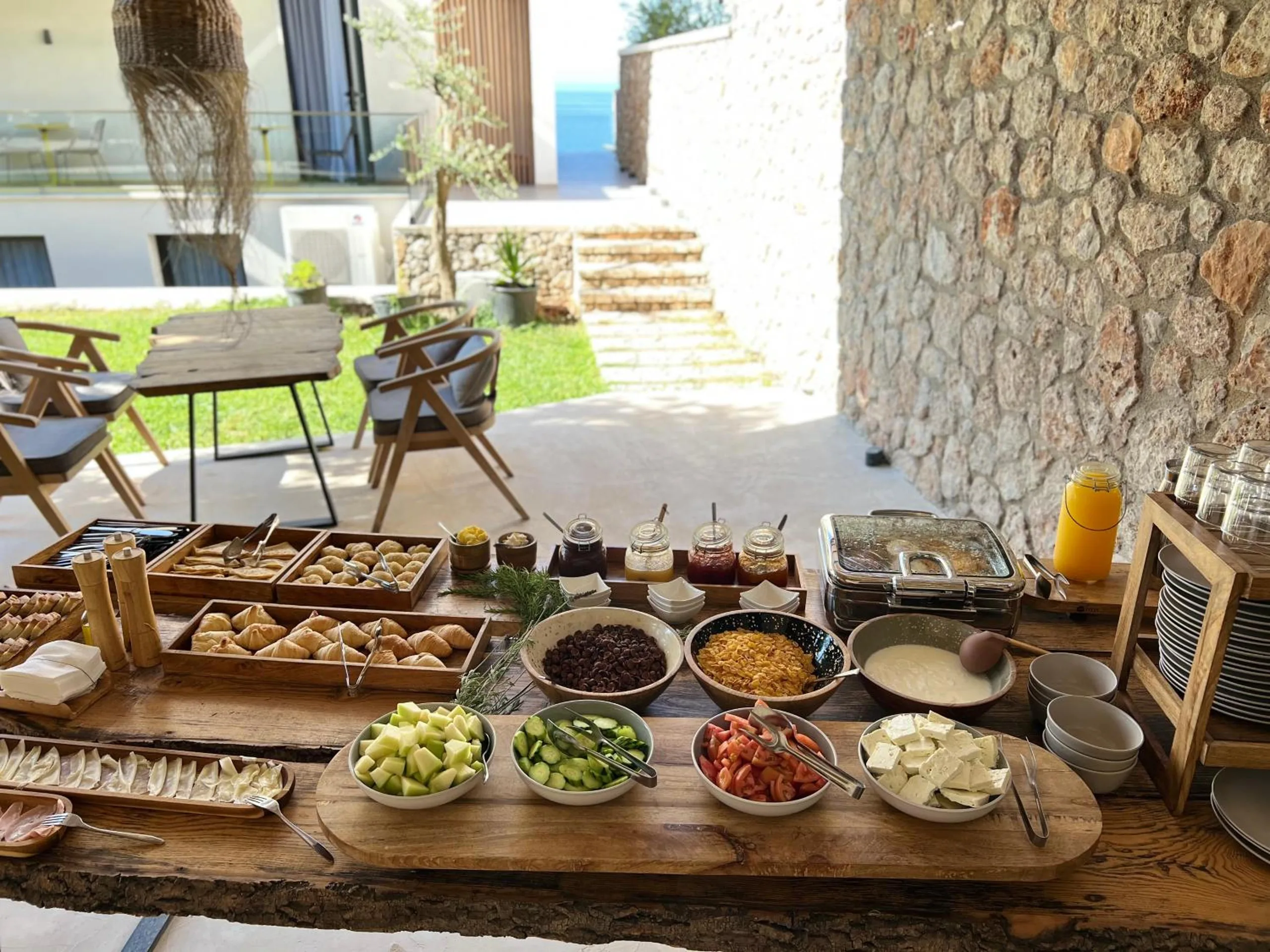 A breakfast buffet laid along a live-edge board on the terrace — bowls of cucumber, tomato, cheese, cereals and olives, trays of pastries and jars of jam — with the lawn and the house behind