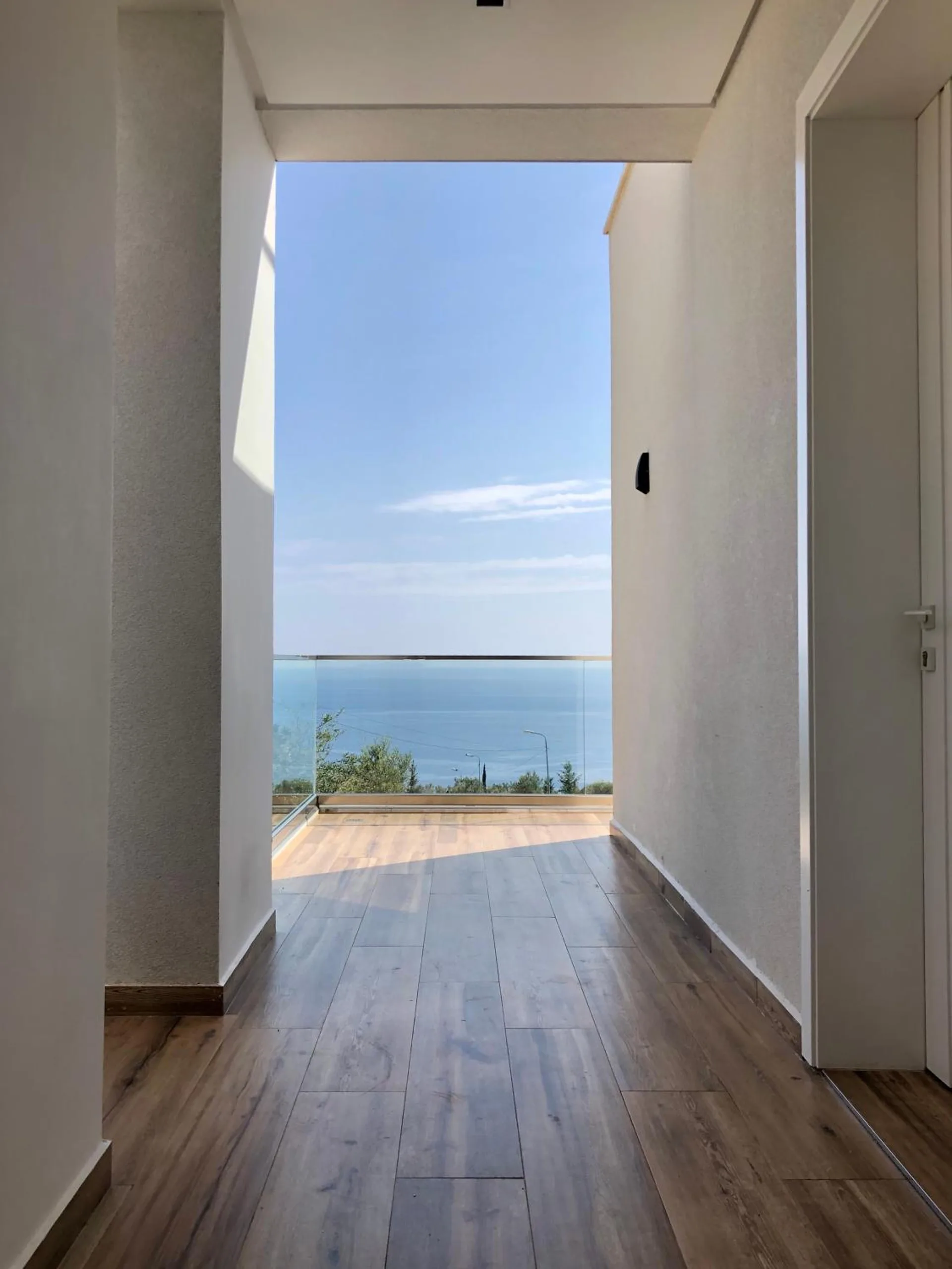 A white corridor with a timber floor ending in an open doorway onto a terrace, and the sea filling the doorway