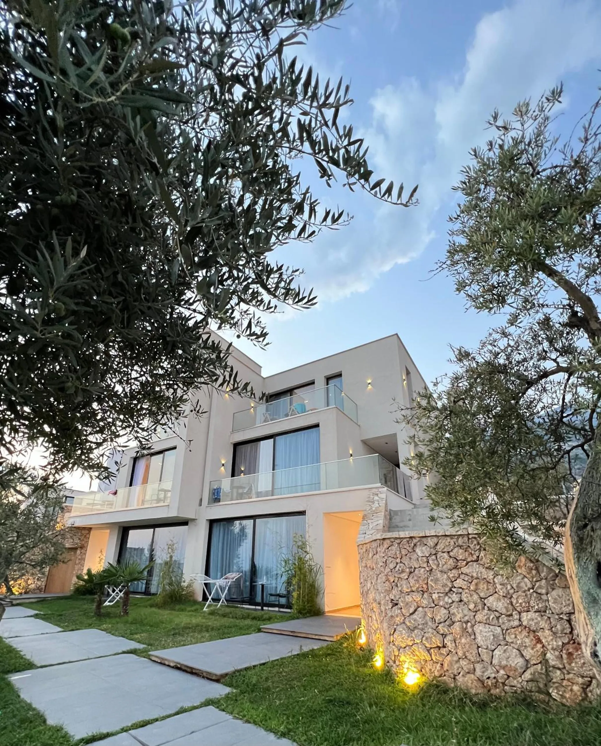 The hotel's greige render and black-framed windows seen between two olive trees, with a lit dry-stone wall beside the garden path
