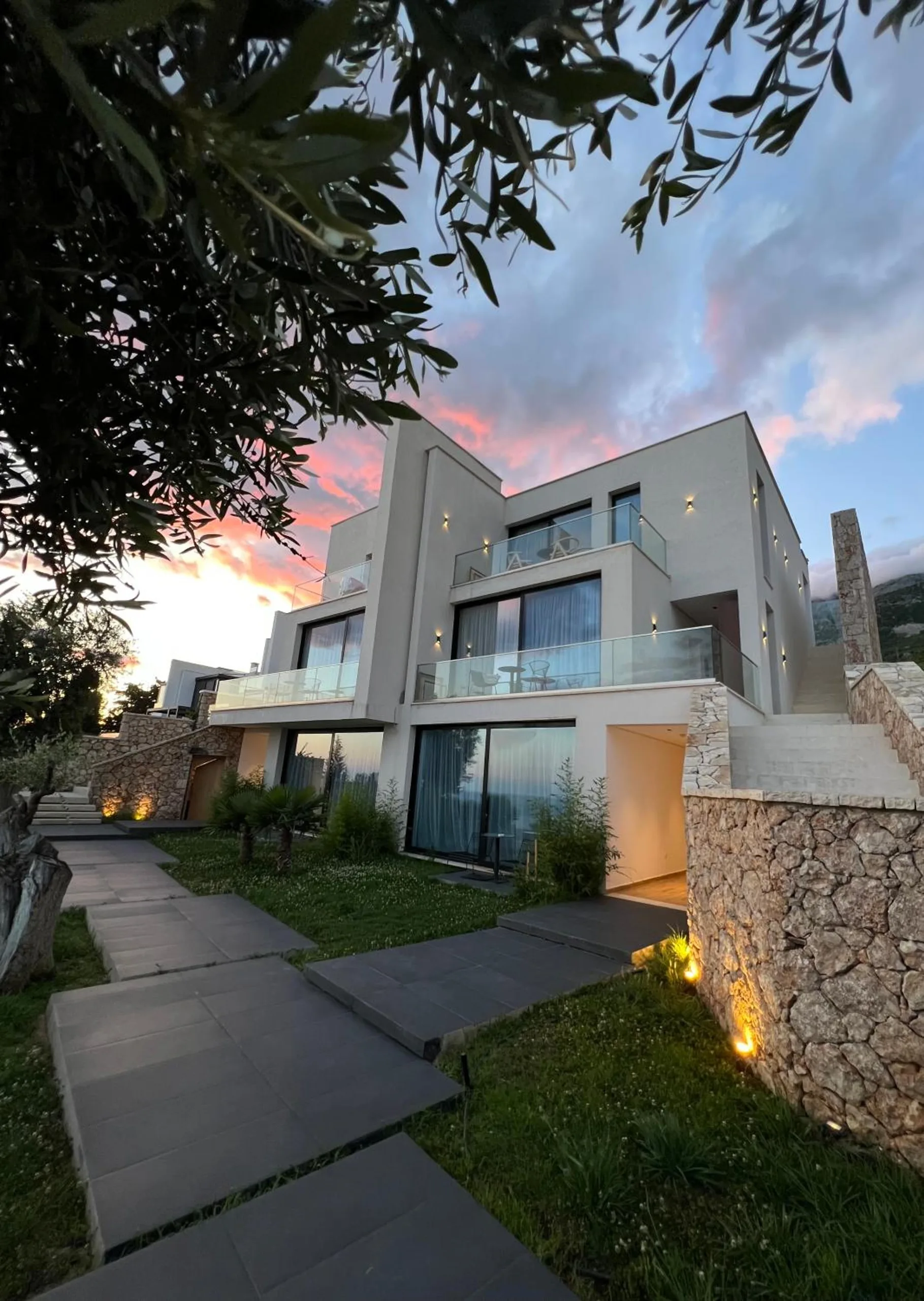 The three-storey hotel at dusk under a pink sky, its glass balconies lit, a stone retaining wall and a paved path through the lawn in the foreground