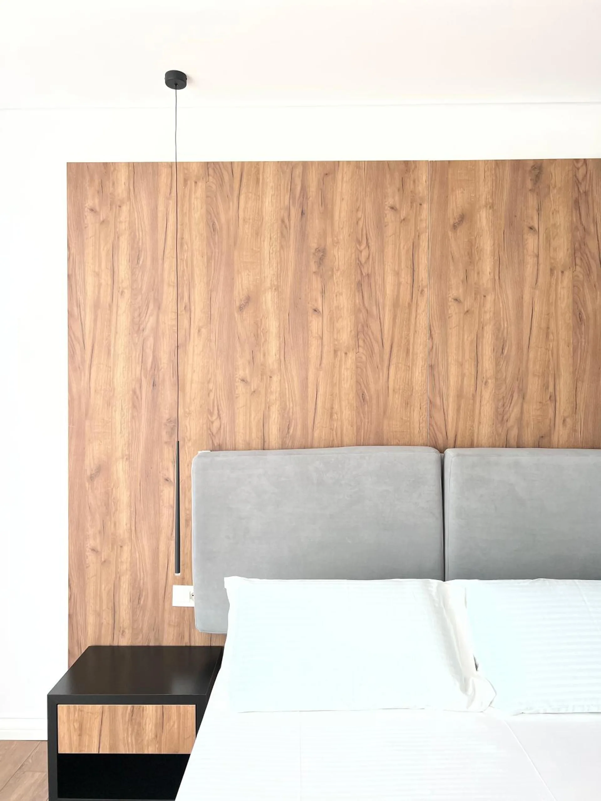A grey upholstered headboard set into a walnut-veneer wall above a white bed, with a black bedside table and a pendant lamp