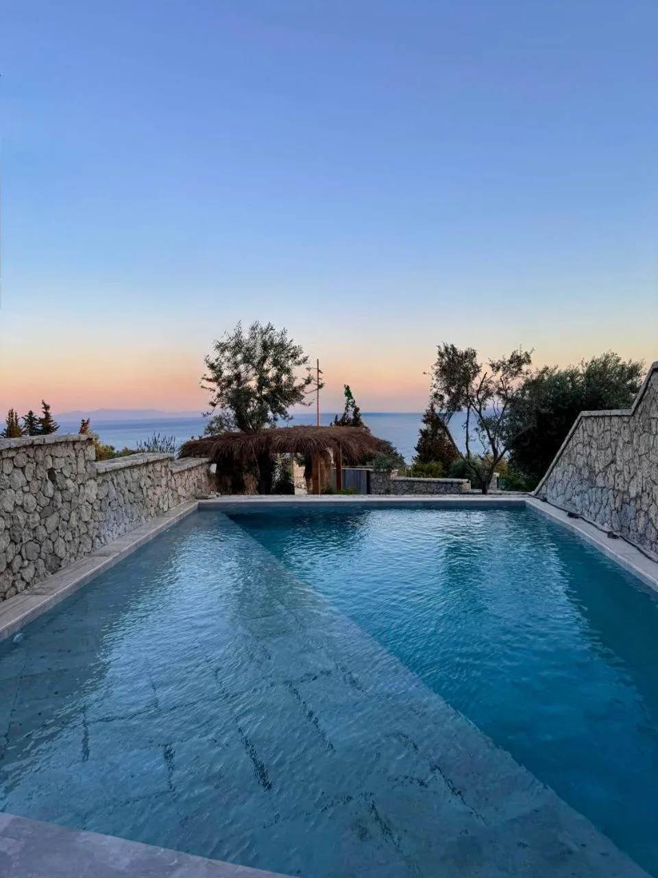 The pool at sunset between dry-stone walls, olive trees and a reed-roofed shelter at the far end, the sea and a pink horizon beyond