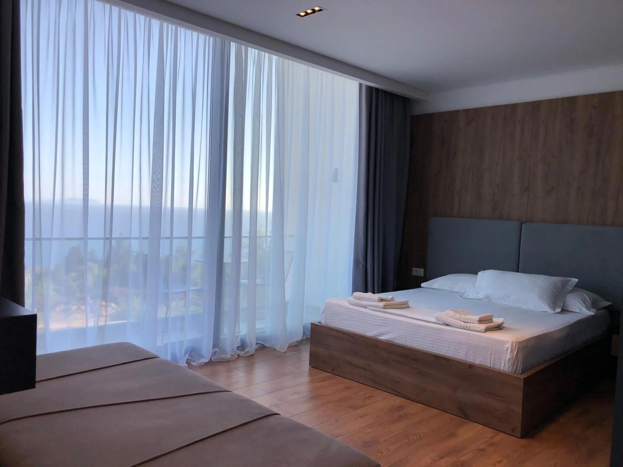 A double bed with a grey headboard and walnut wall beside a tall window, the sea and the balcony seen through the sheer