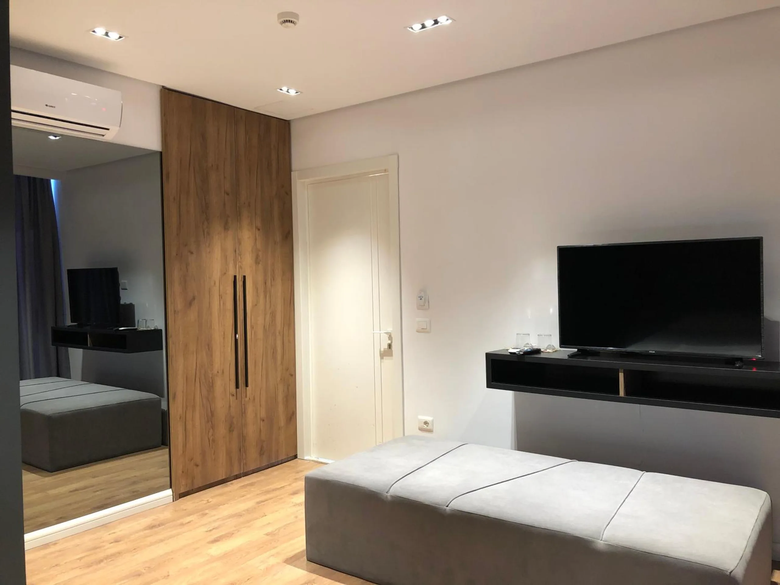 A grey upholstered bench, a black wall-hung desk with a television above it, a walnut wardrobe and a mirror reflecting the room