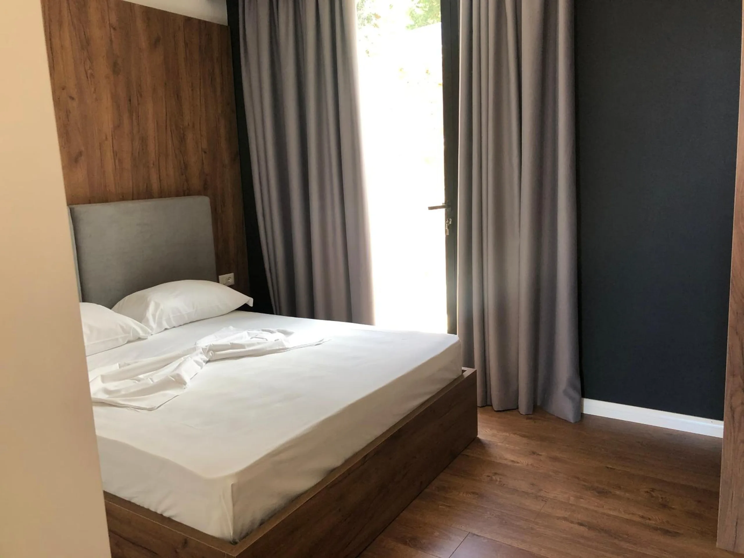 A double bed with a grey upholstered headboard against a walnut panel and a black wall, grey curtains drawn back from a bright doorway