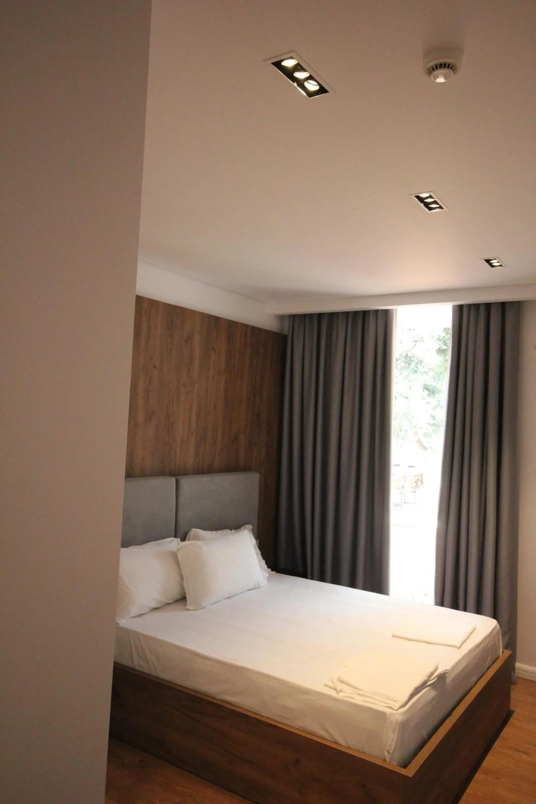 A double bed with a grey headboard and a walnut wall beside a tall window with grey curtains