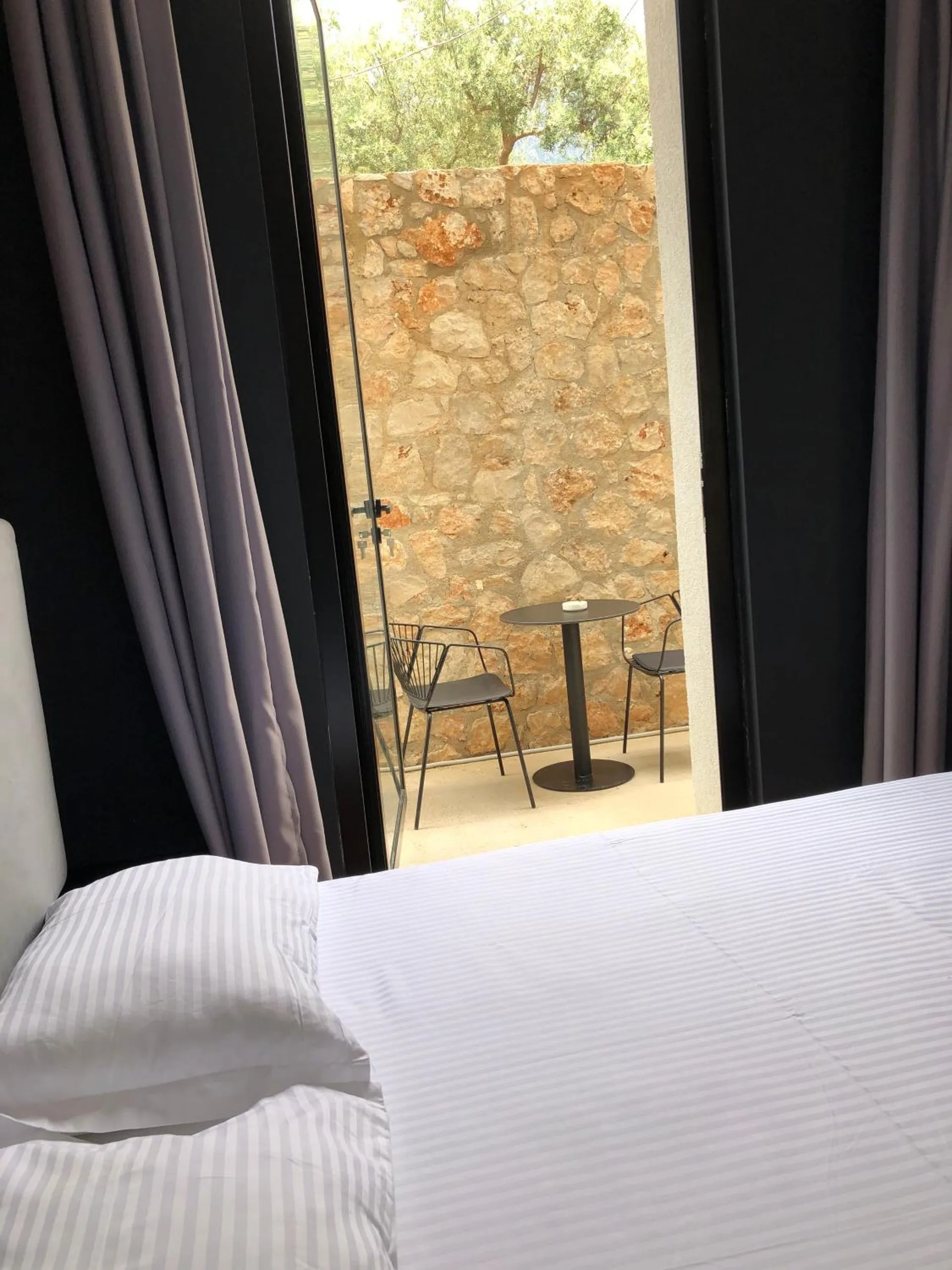 The corner of a white-made bed by a tall window, and outside a small paved patio with a bistro table and chairs against a dry-stone wall