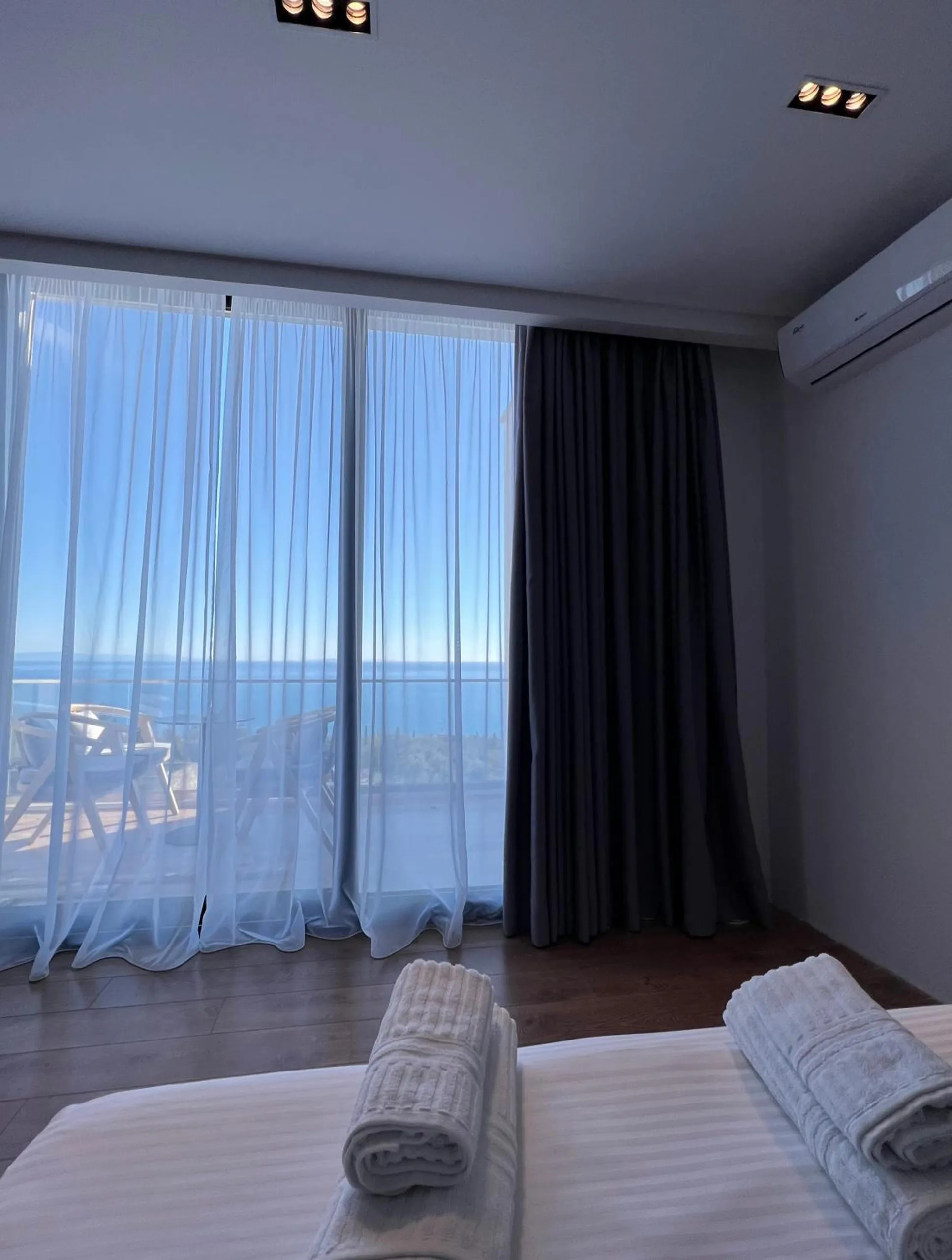 Rolled towels on a striped double bed facing a glass wall with white sheers, a balcony and the open sea beyond