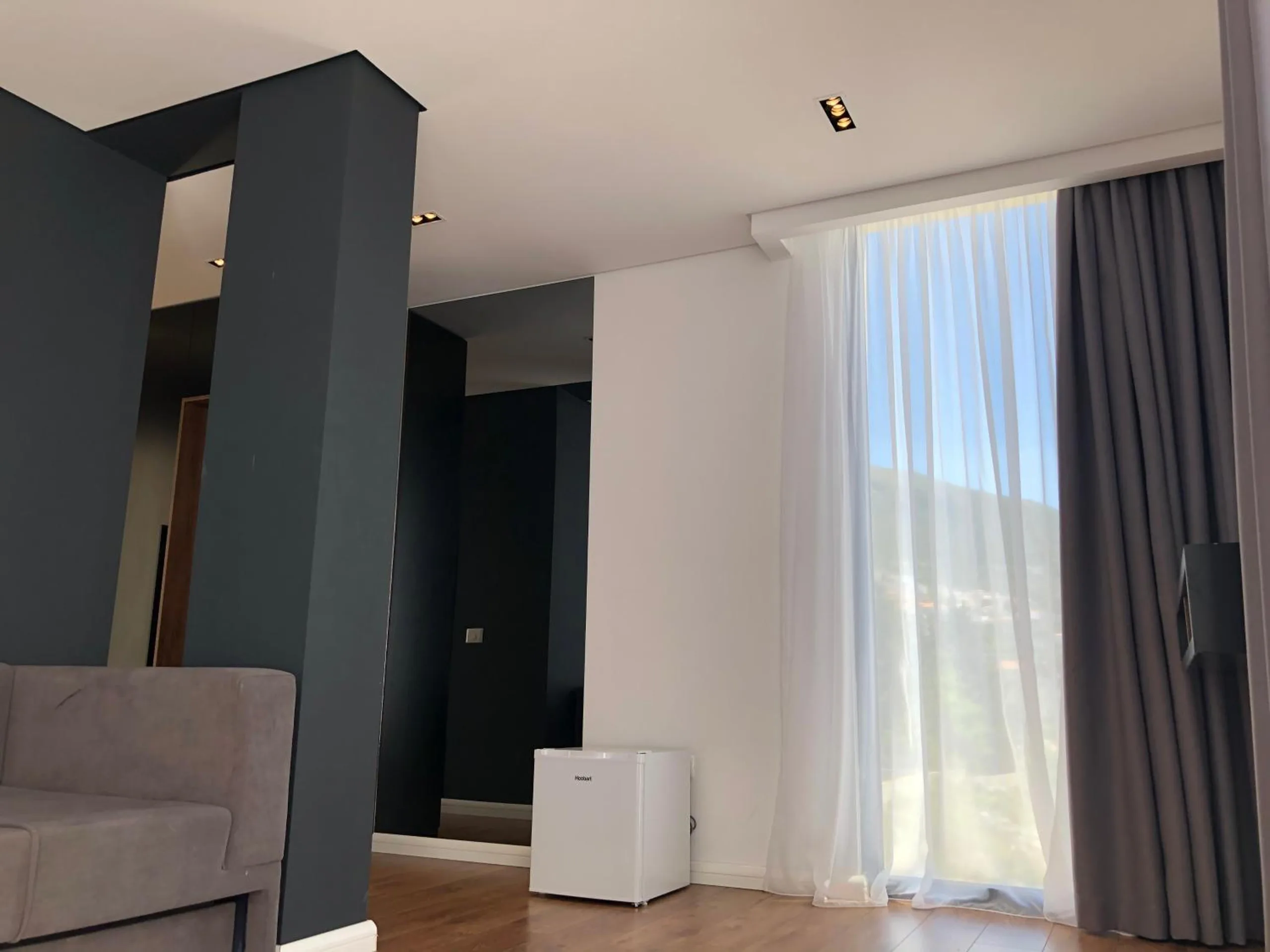 A graphite pillar and a minibar beside a tall window with a white sheer and a grey curtain, green hills outside