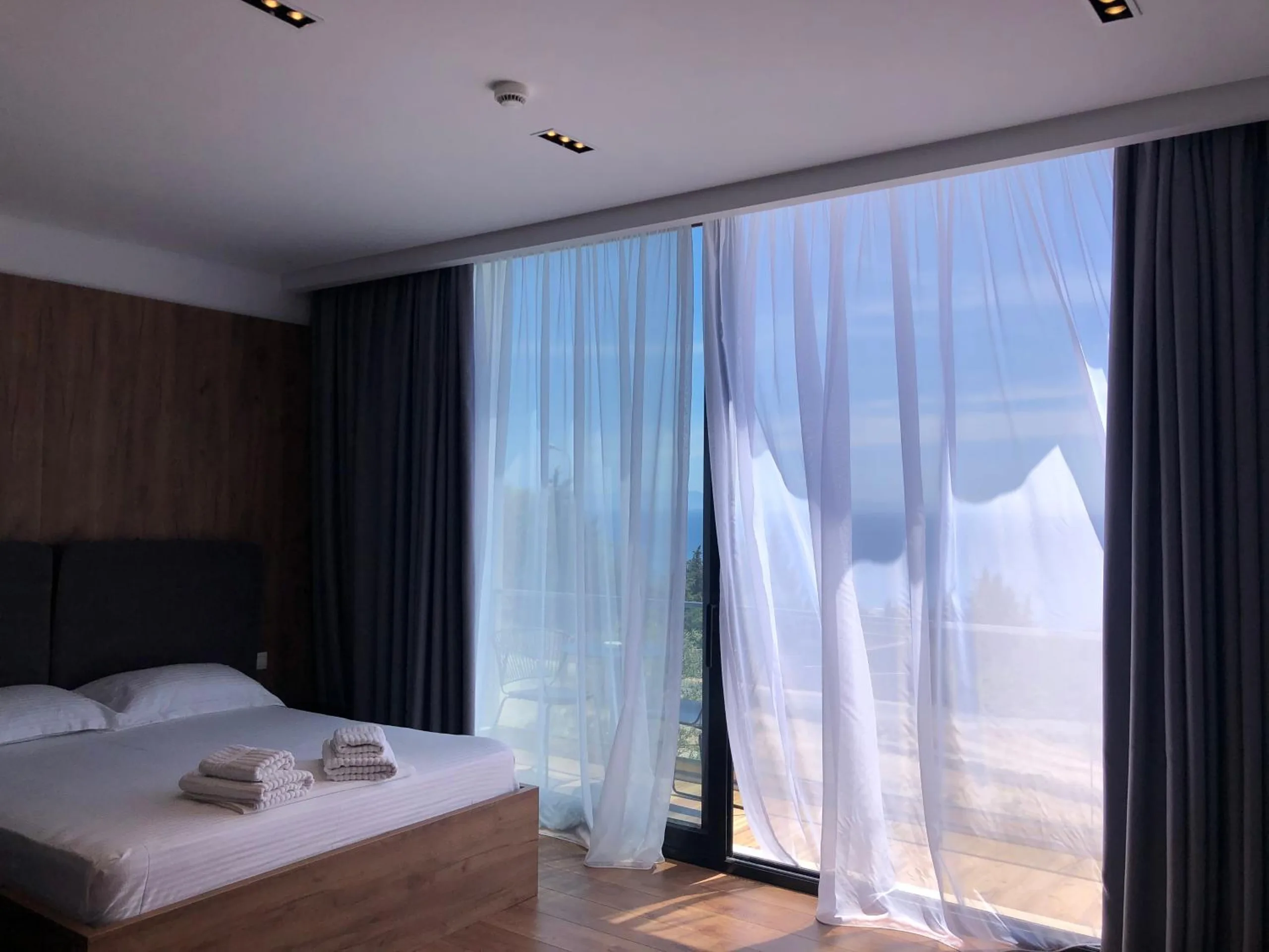 A double bed against a walnut wall, and beyond it a wall of glass hung with white sheers blowing in from an open balcony, the sea pale blue behind them