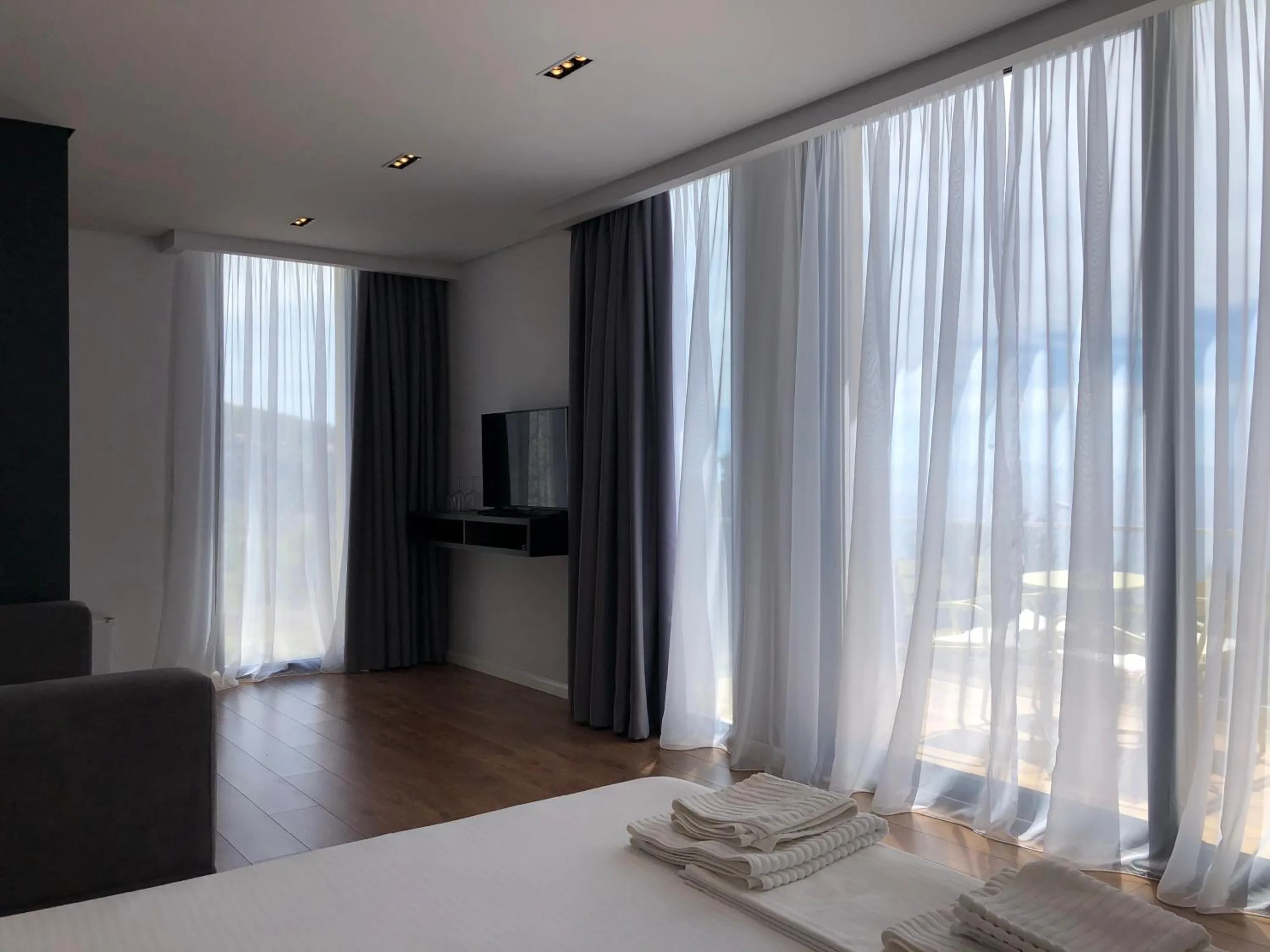 A wide bright room with floor-to-ceiling white sheers and grey curtains along a glass wall, towels folded on the bed in the foreground