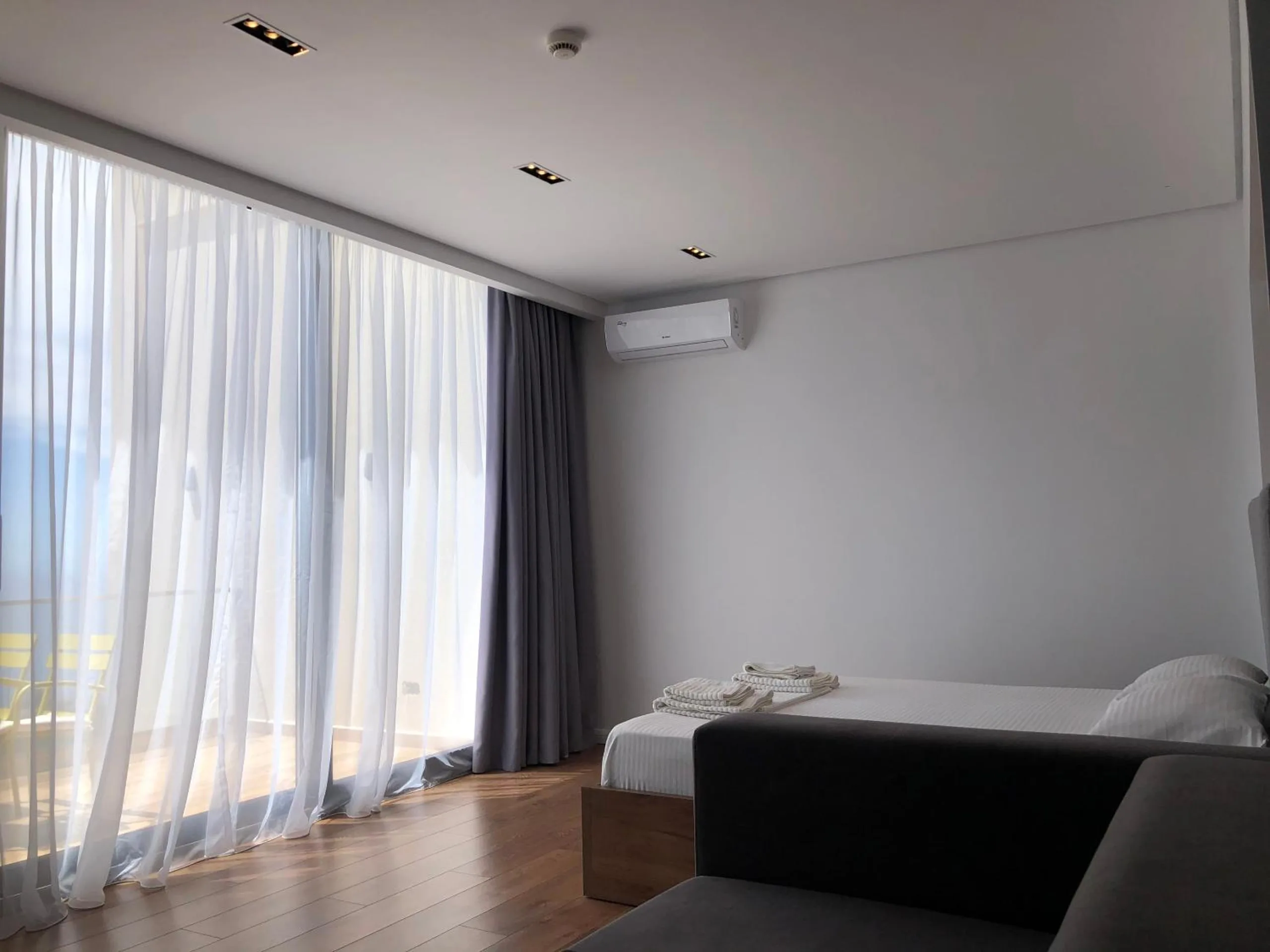 A grey sofa and a bed end below a glass wall hung with white sheers, an air-conditioning unit high on the white wall
