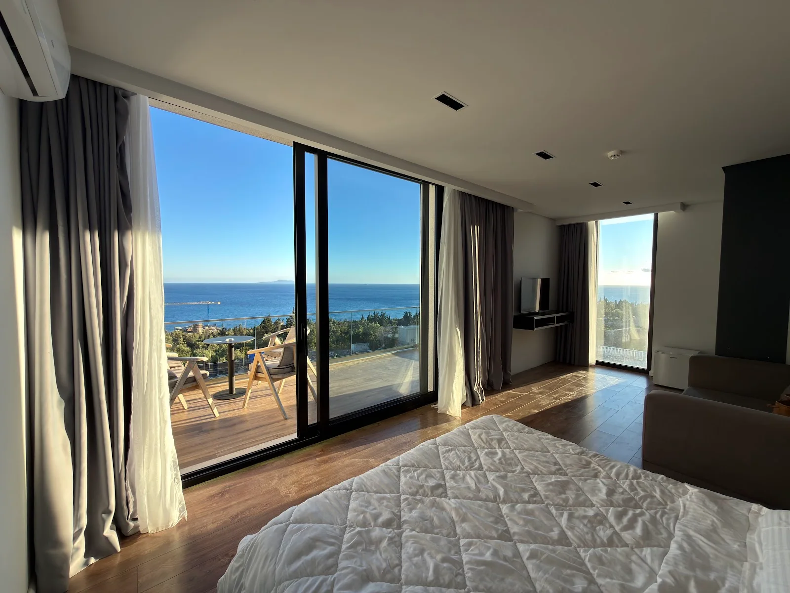 A large room with a quilted double bed and a sofa, its sliding glass doors open onto a timber terrace with chairs, the bay and the open sea beyond