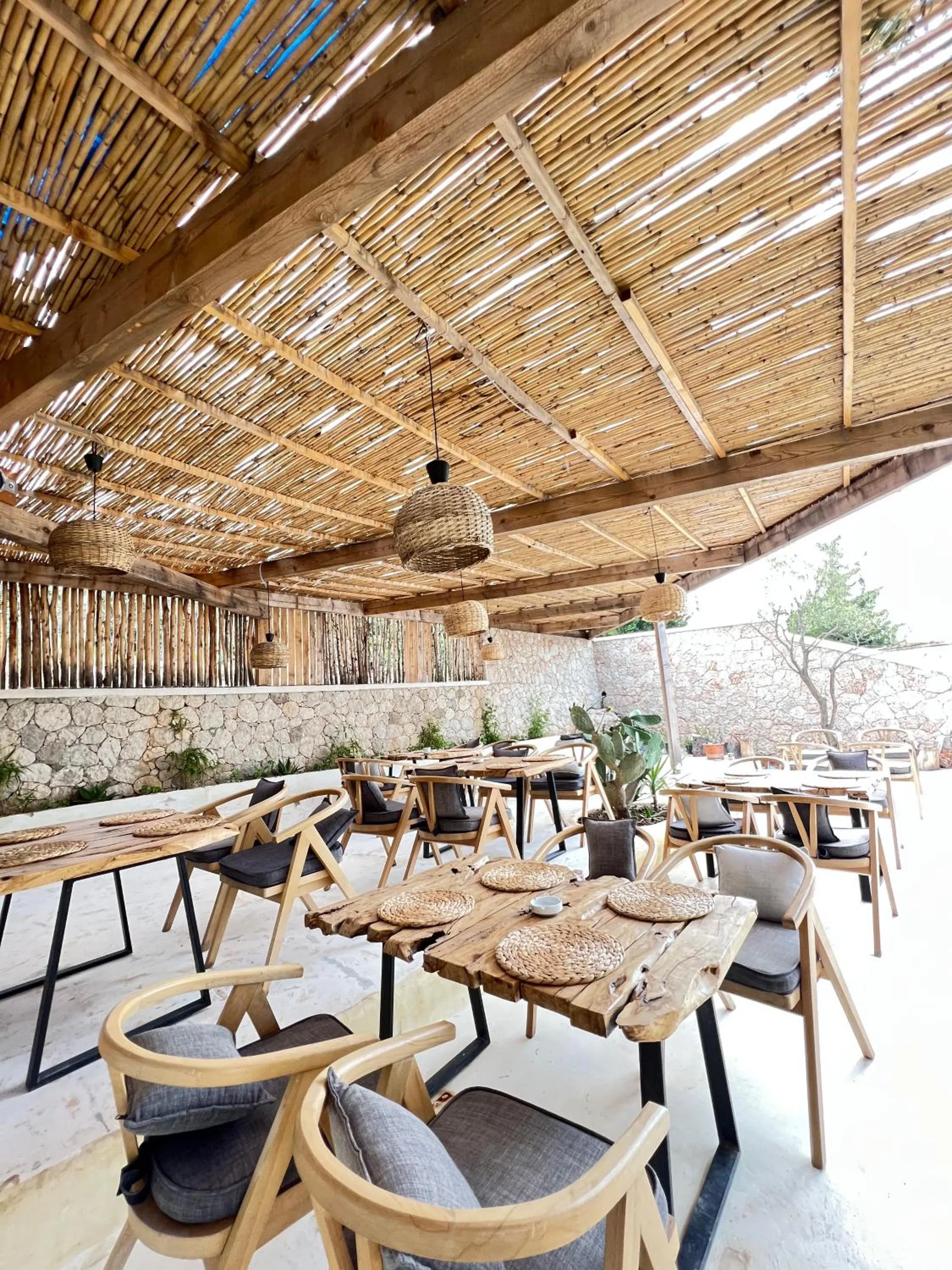 Long timber tables set out under the reed pergola, a woven pendant lamp hanging over them and a dry-stone wall at the back