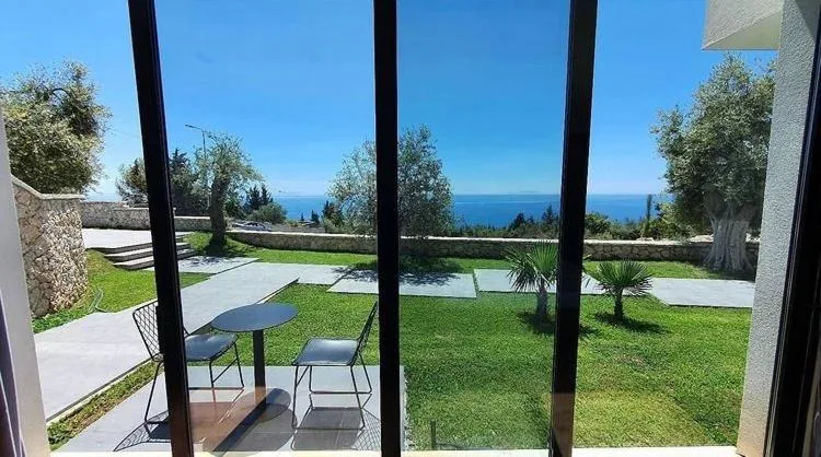 The lawn, young palms and olive trees seen through a black-framed glass wall, the sea along the horizon