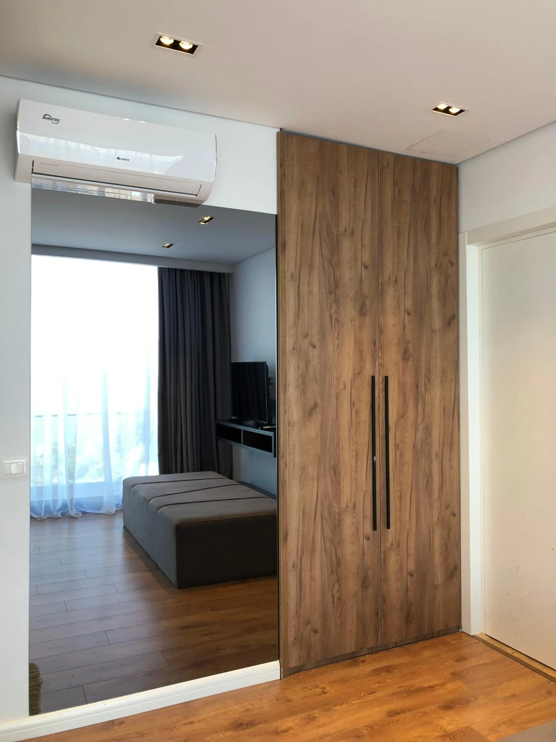 A tall walnut wardrobe with long black handles beside a full-length mirror that reflects the window and the bed