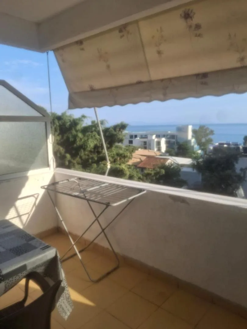 A tiled balcony with a clothes airer and a table, its awning open, looking over trees and rooftops to the sea