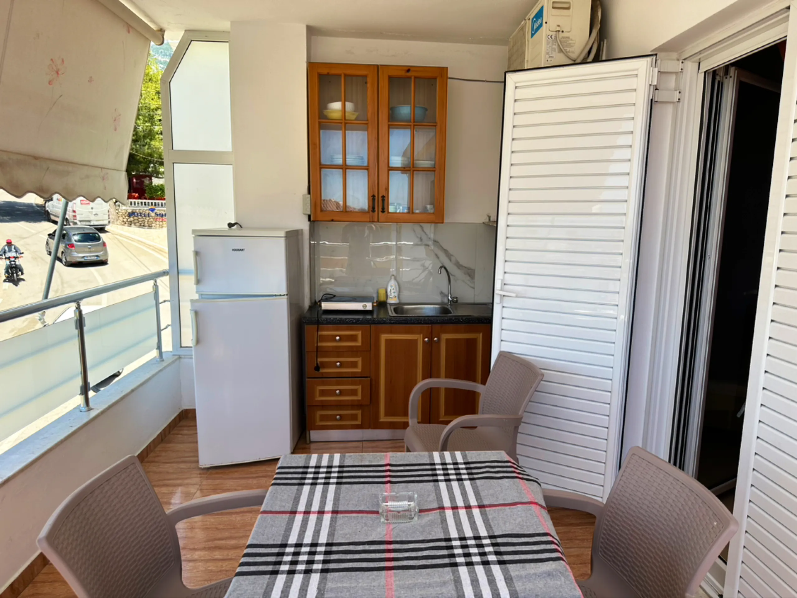 A kitchenette on a covered balcony: glass-fronted cupboard, sink, two-ring hob and fridge, with a table and three chairs and the street below