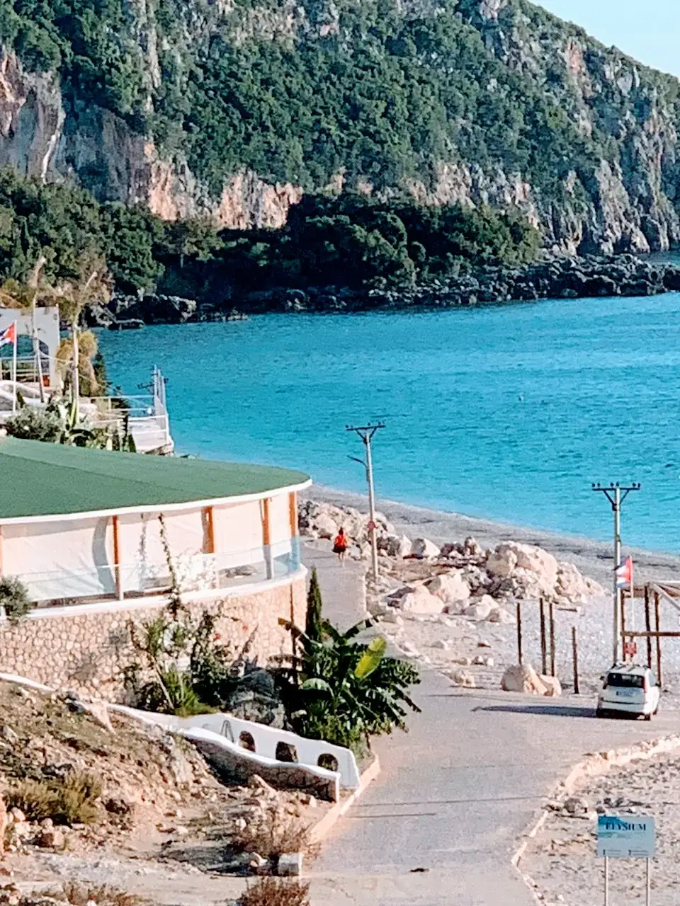 A paved lane running down past a beach club to a white pebble shore and turquoise water under wooded limestone cliffs