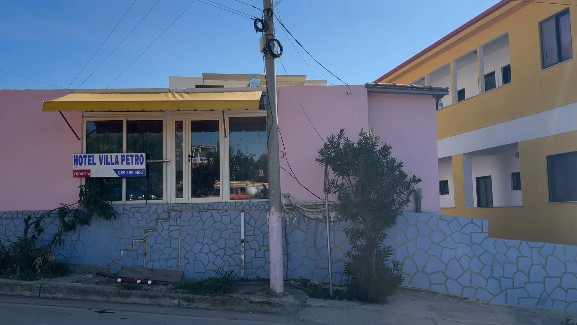 A pink single-storey front with a yellow scalloped awning above a periwinkle crazy-paving wall, beside a yellow two-storey block, seen from the road