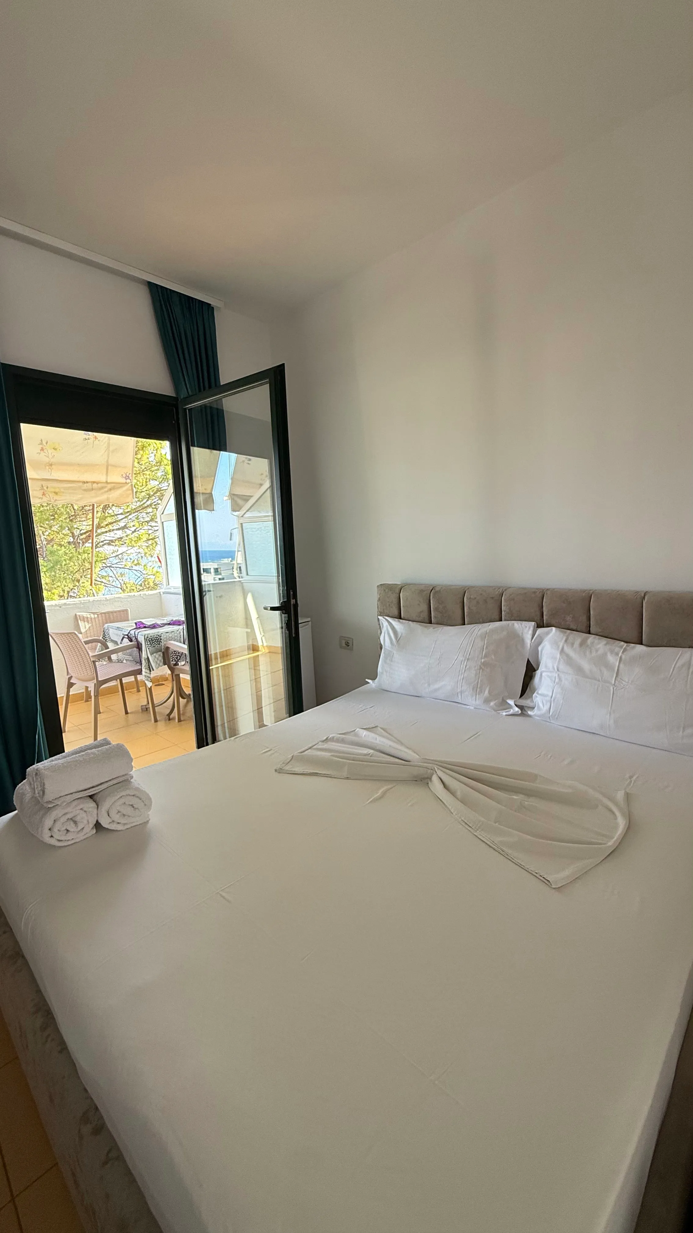 A made-up double bed with rolled towels, and an open aluminium door onto a balcony with a table, chairs and a strip of sea beyond the pines