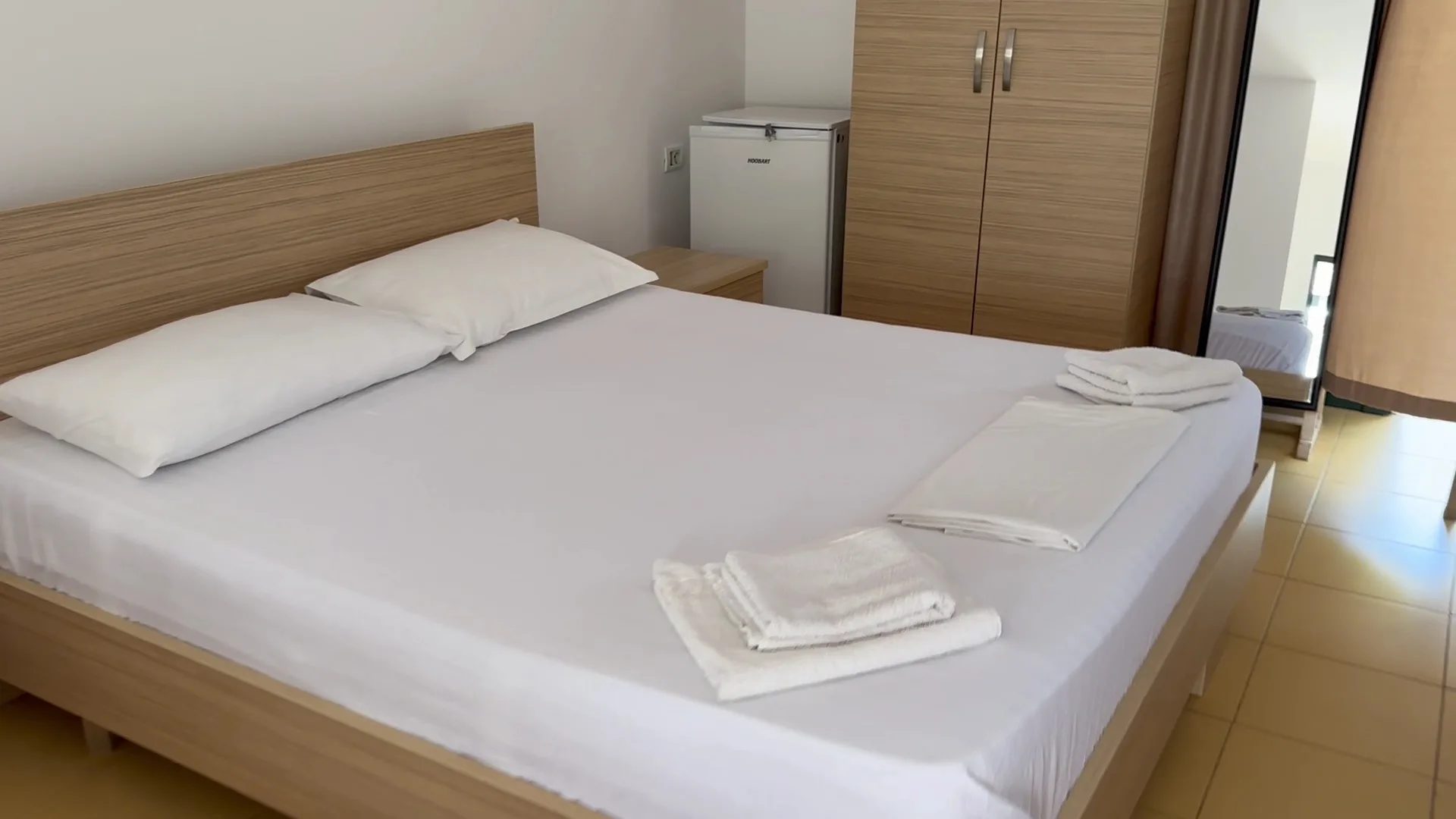 A double bed in white linen with folded towels, a pale oak wardrobe, a small fridge and a mirror on a tiled floor