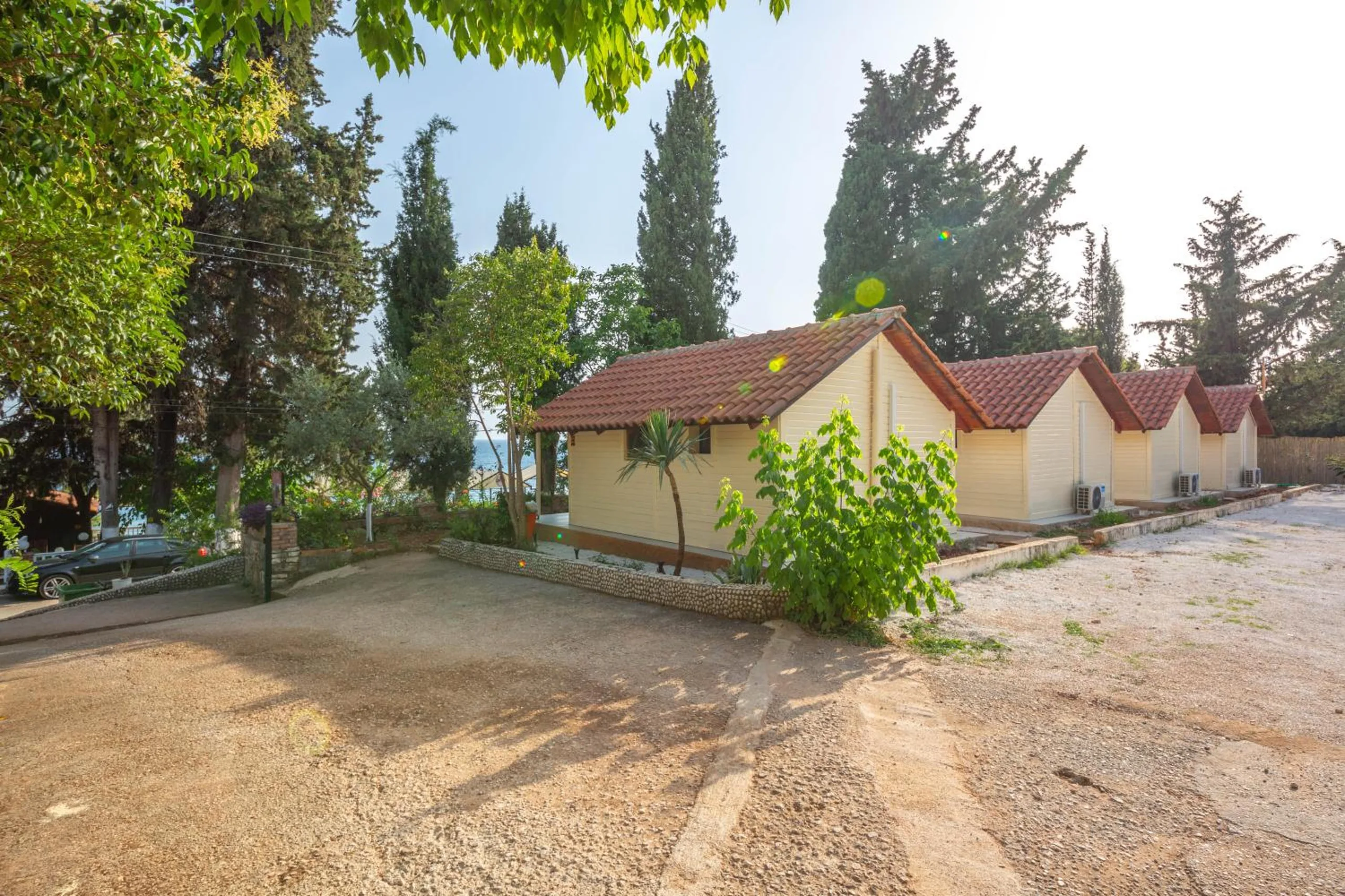 A row of cream timber bungalows with terracotta pantile roofs under cypress trees, a gravel yard in front