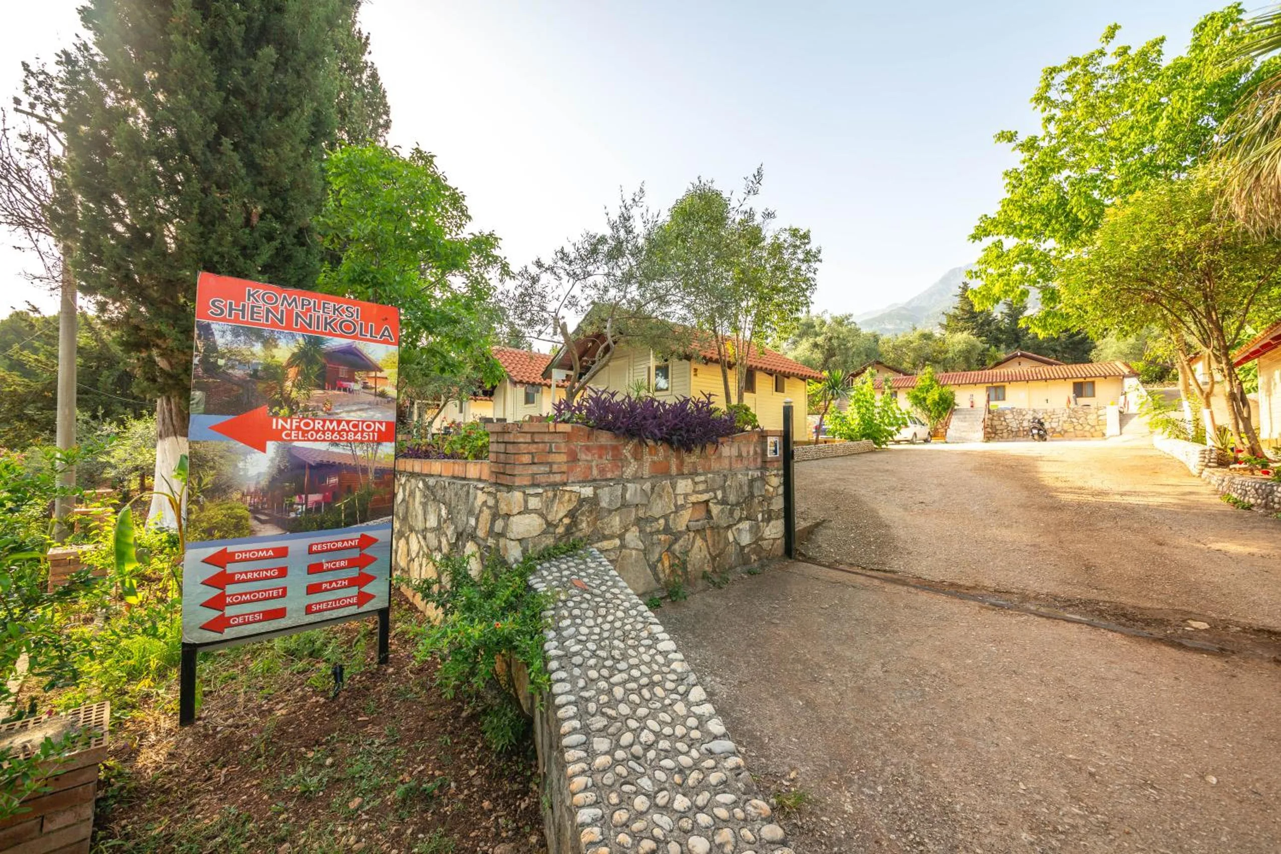 The entrance drive with the property's red and blue sign reading Kompleksi Shen Nikolla, red arrows pointing to the rooms, parking, restaurant, pizzeria and beach