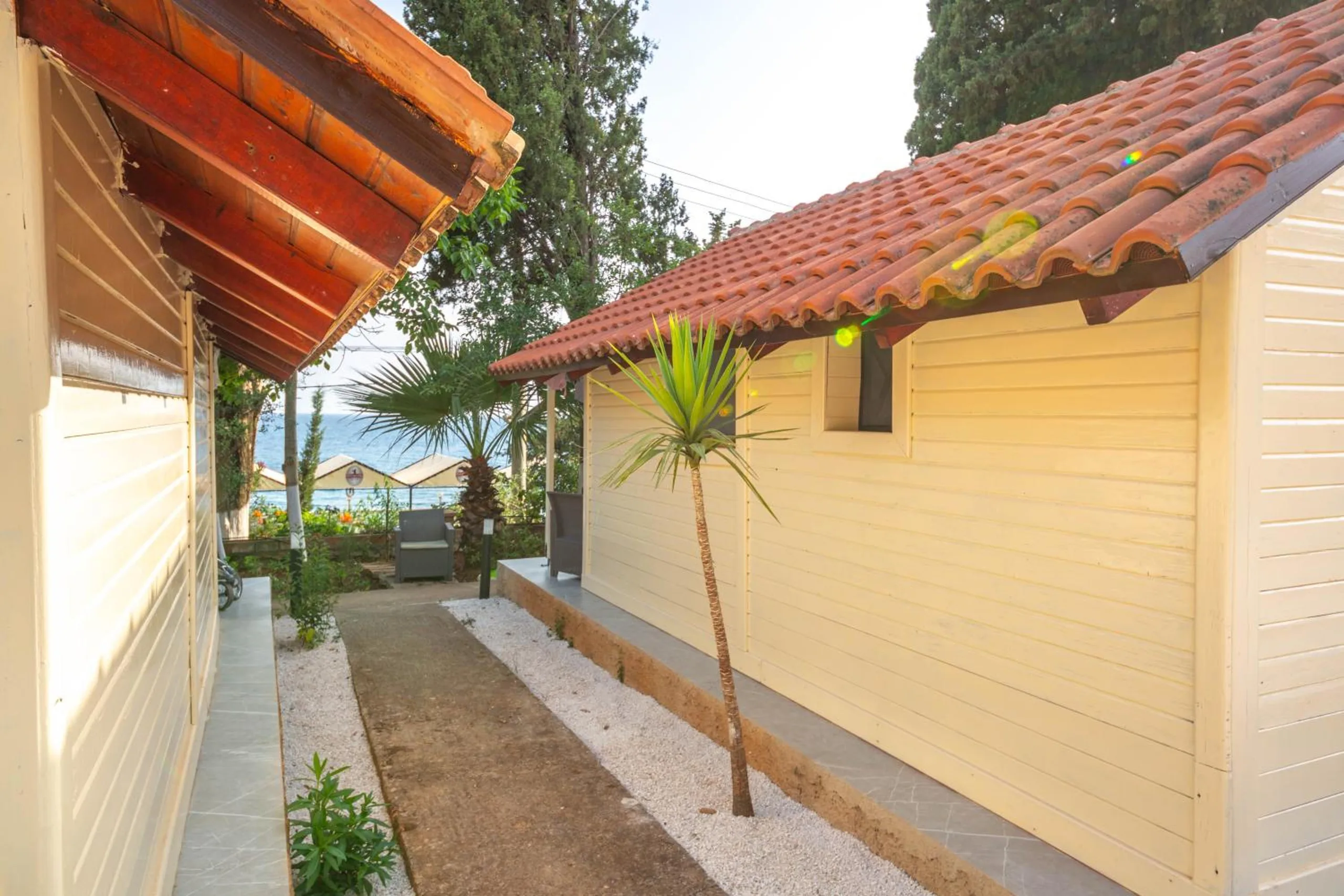 A gravel path between two cream timber bungalows, a young palm against one wall, garden chairs at the end and the sea and beach umbrellas beyond