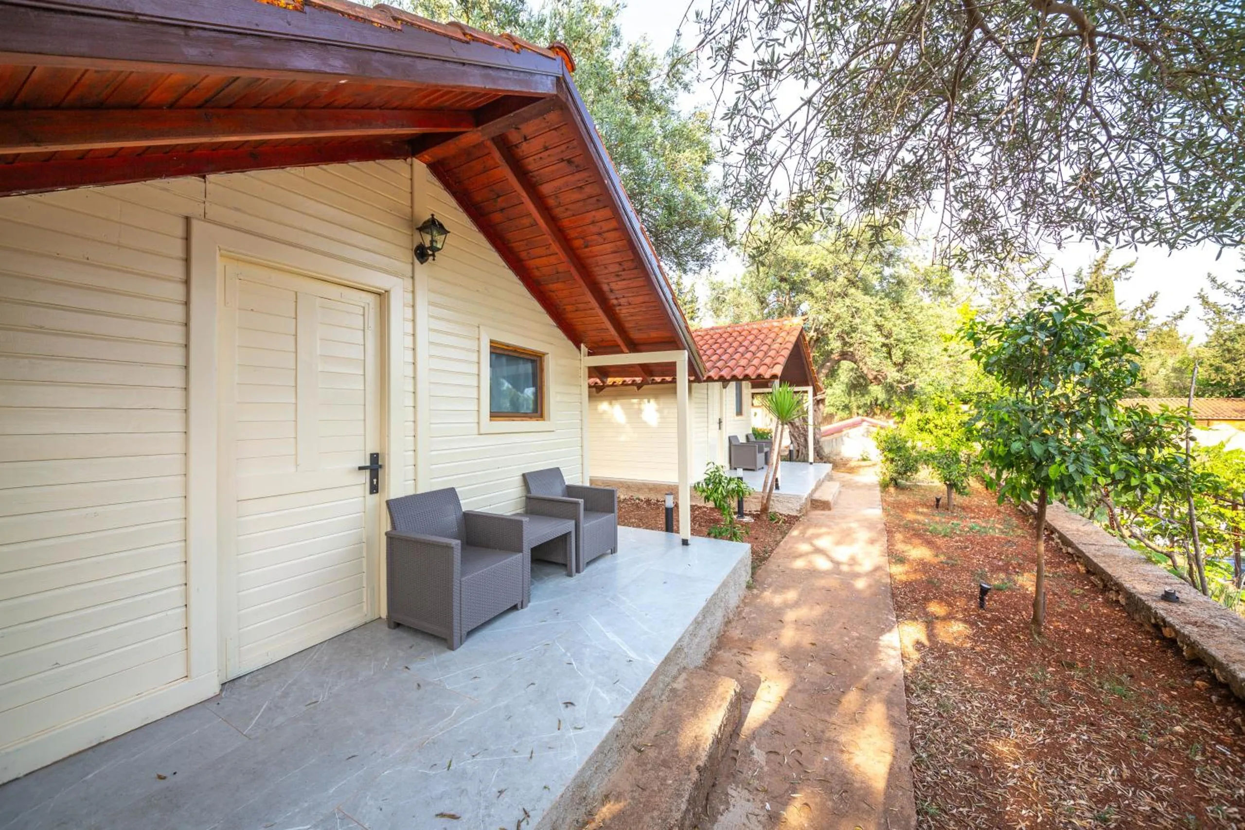 The covered porch of a cream bungalow with two grey wicker armchairs and a small table beside the door, a garden path running past