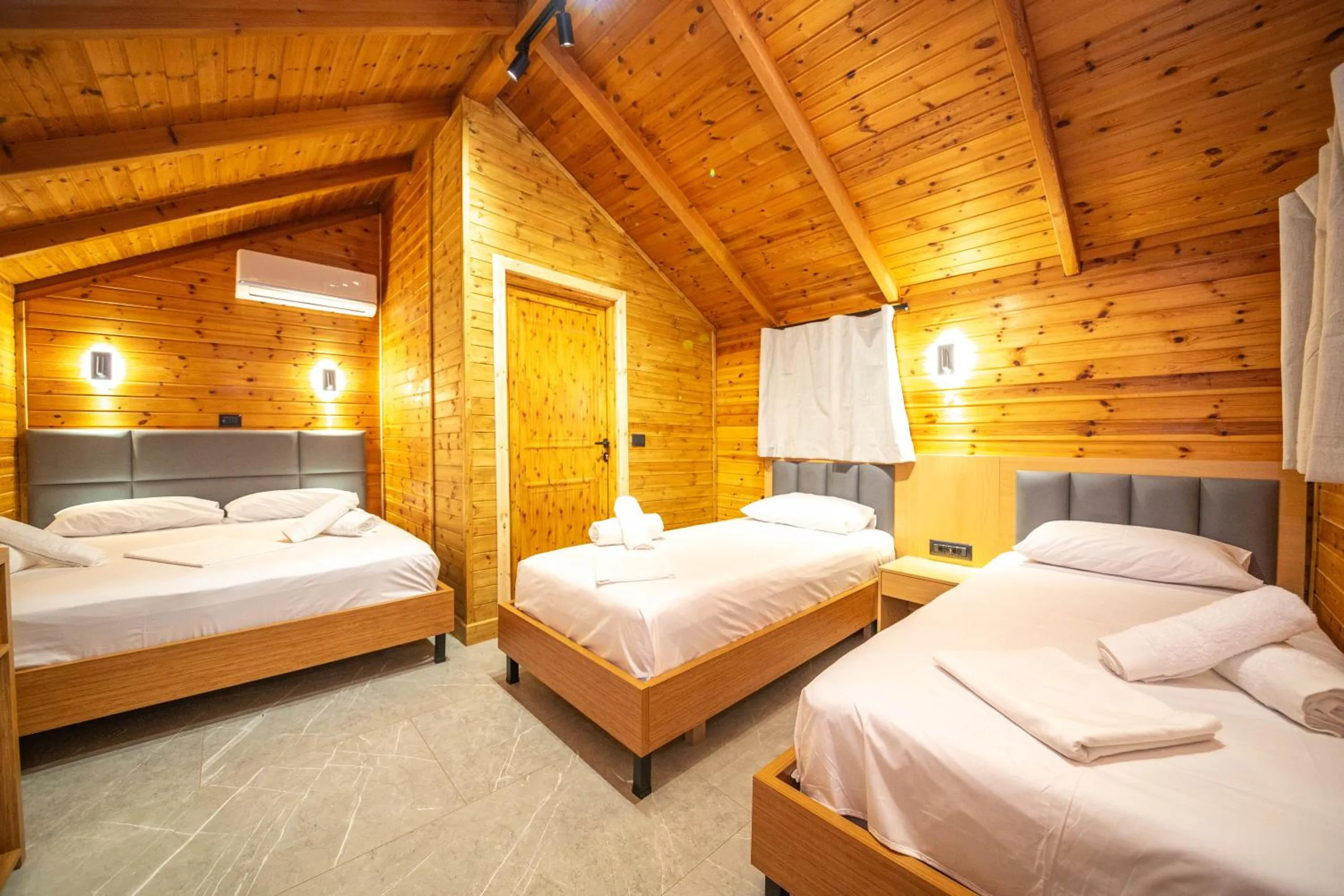 Inside a bungalow: knotty-pine walls and a pitched pine ceiling, a queen bed and two single beds with grey upholstered headboards, wall lights and an air-conditioning unit